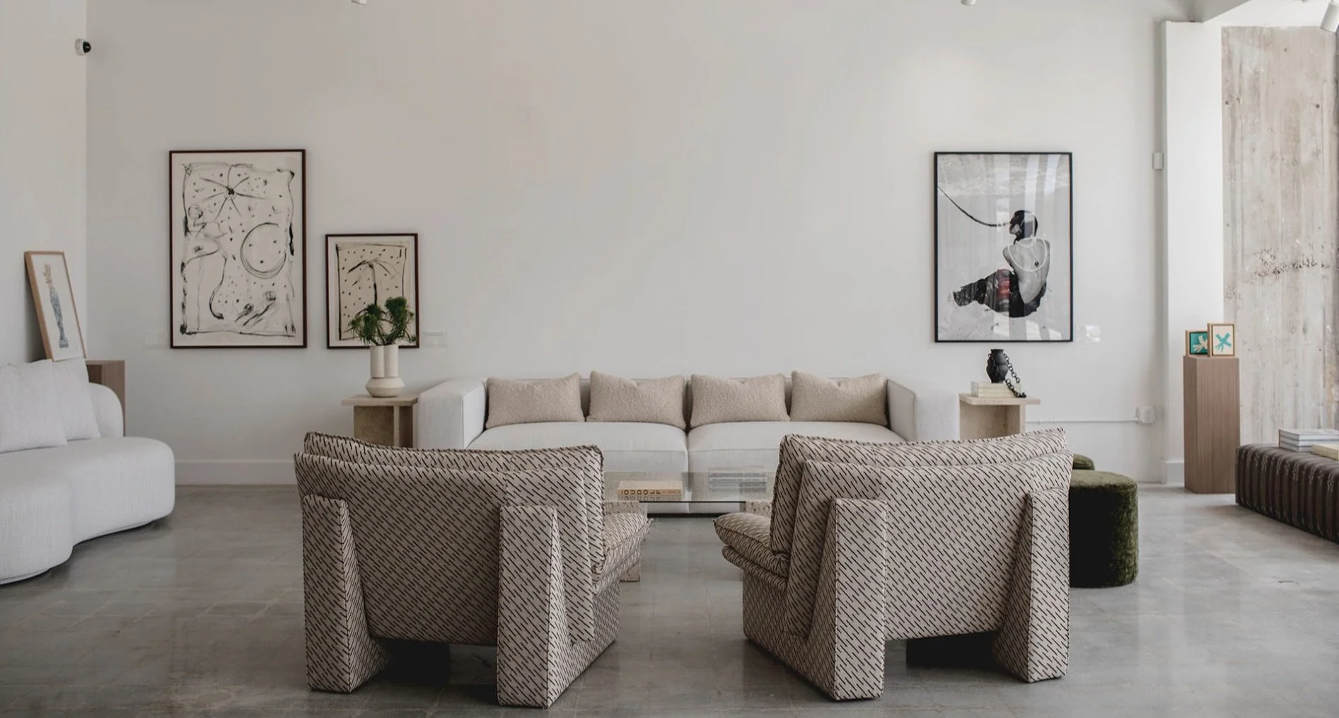 Contemporary living room with white walls, multiple framed artwork, and a variety of seating including a white sofa, patterned armchairs, and a bench. Coffee table, potted plants, and decorative objects are present.