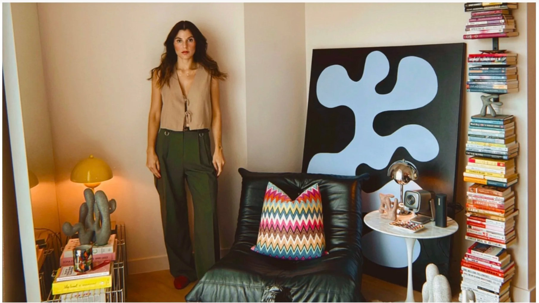 A woman with brown hair wearing a beige sleeveless vest and dark green pants standing in a living room next to a black leather chair with a colorful zigzag pillow. The room has a modern art piece with black and white shapes on the wall, a side table with a metallic lamp and decorative items, a tall bookshelf filled with books, and a small cactus sculpture on a wire rack with magazines.