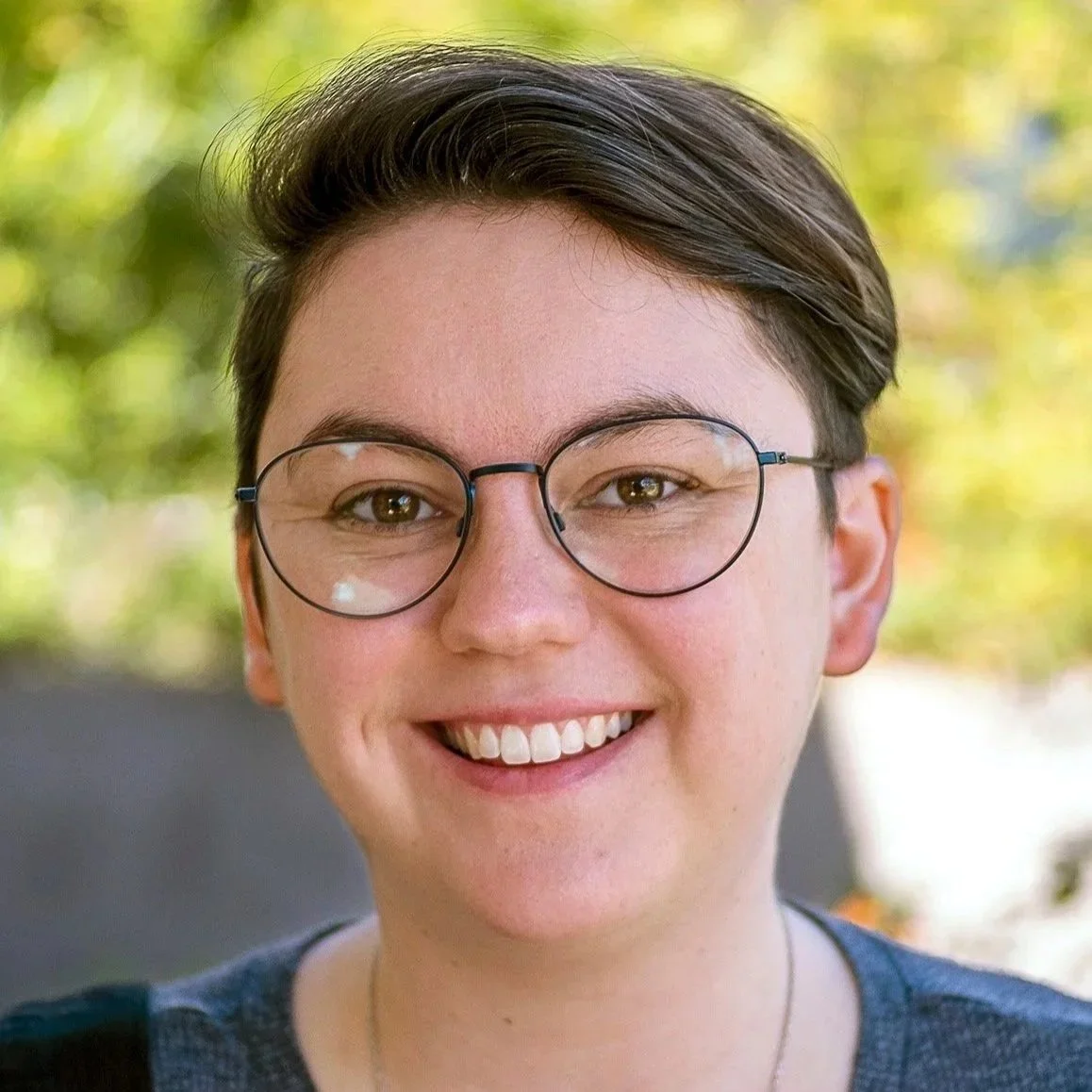 Artist and panelist Bri Noonan smiles squarely at the camera against a green background. Short brown hair and round glasses rest against their fair skin.