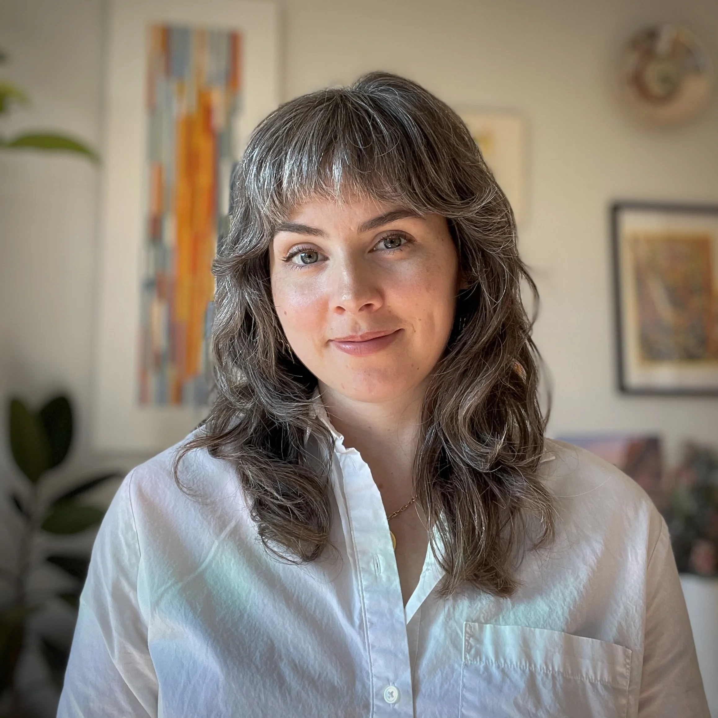 A white woman with long, brown and silver hair and bangs wears a white shirt and smiles at the camera.