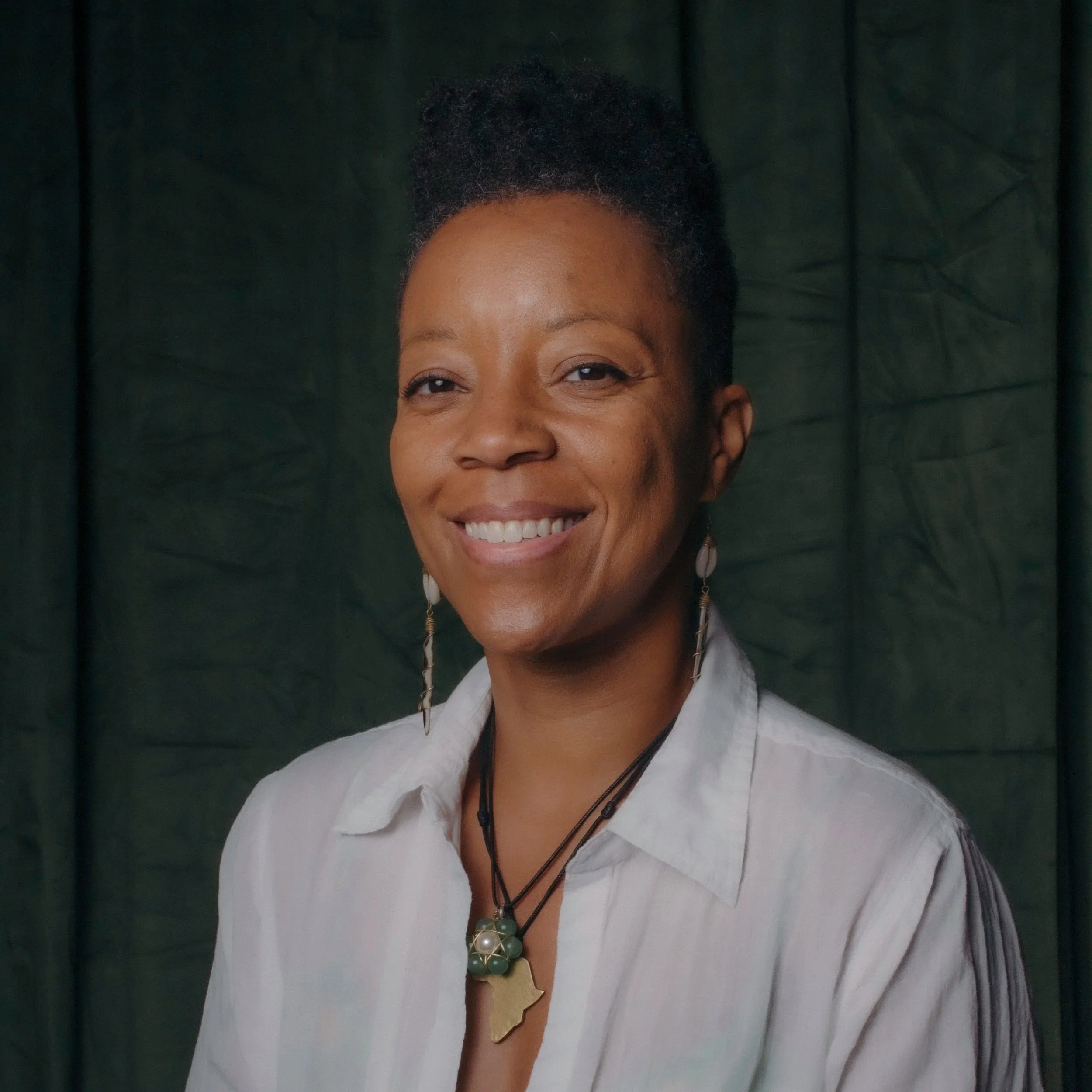 Artist and panelist Tempestt Hazel has warm brown skin, and short black hair. She is smiling against a moss green backdrop in a white button down shirt and delicate gold jewelry.