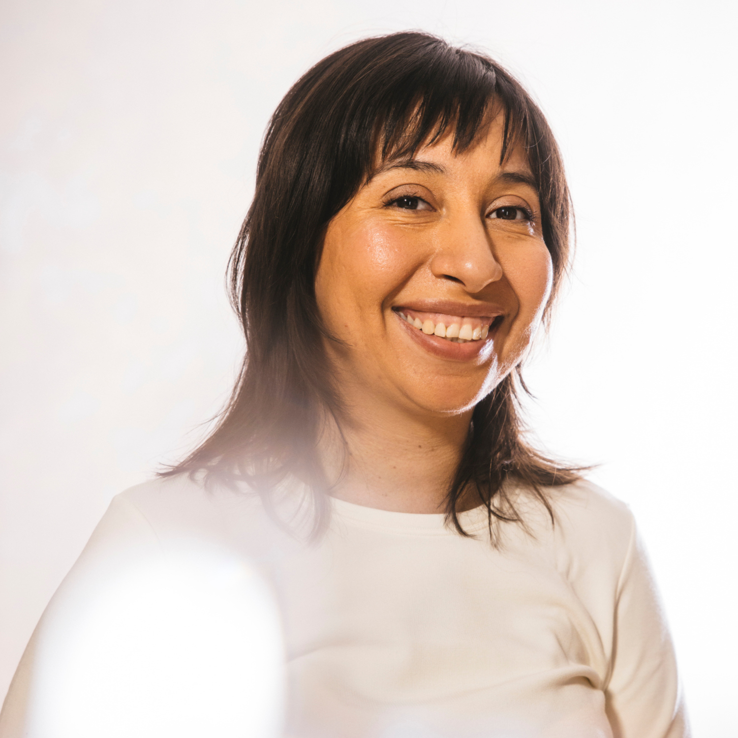 Artist and guest panelist Reveca Torres smiles at the camera against a bright white background. Shoulder length dark brown hair falls around the warm brown skin of her face. She is wearing an off-white long-sleeve shirt.
