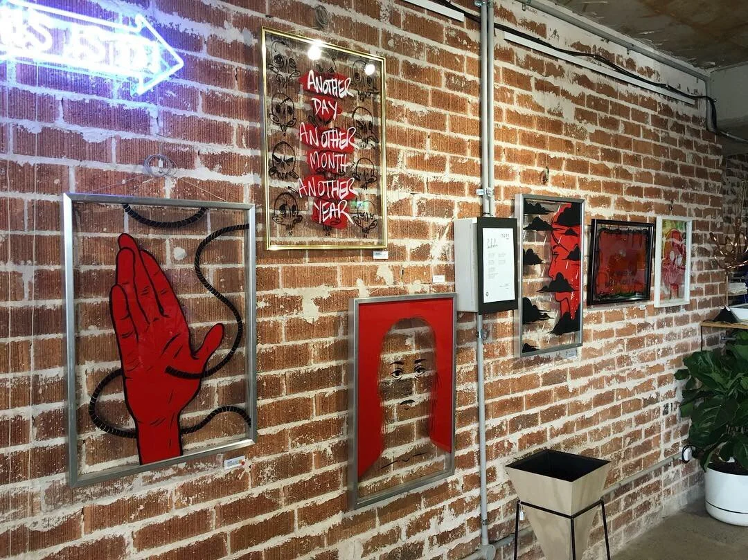 Art gallery wall with various framed artworks, including a red and black hand illustration, a portrait of a woman with red hair, and abstract designs, with a brick wall background and a neon arrow sign pointing left.