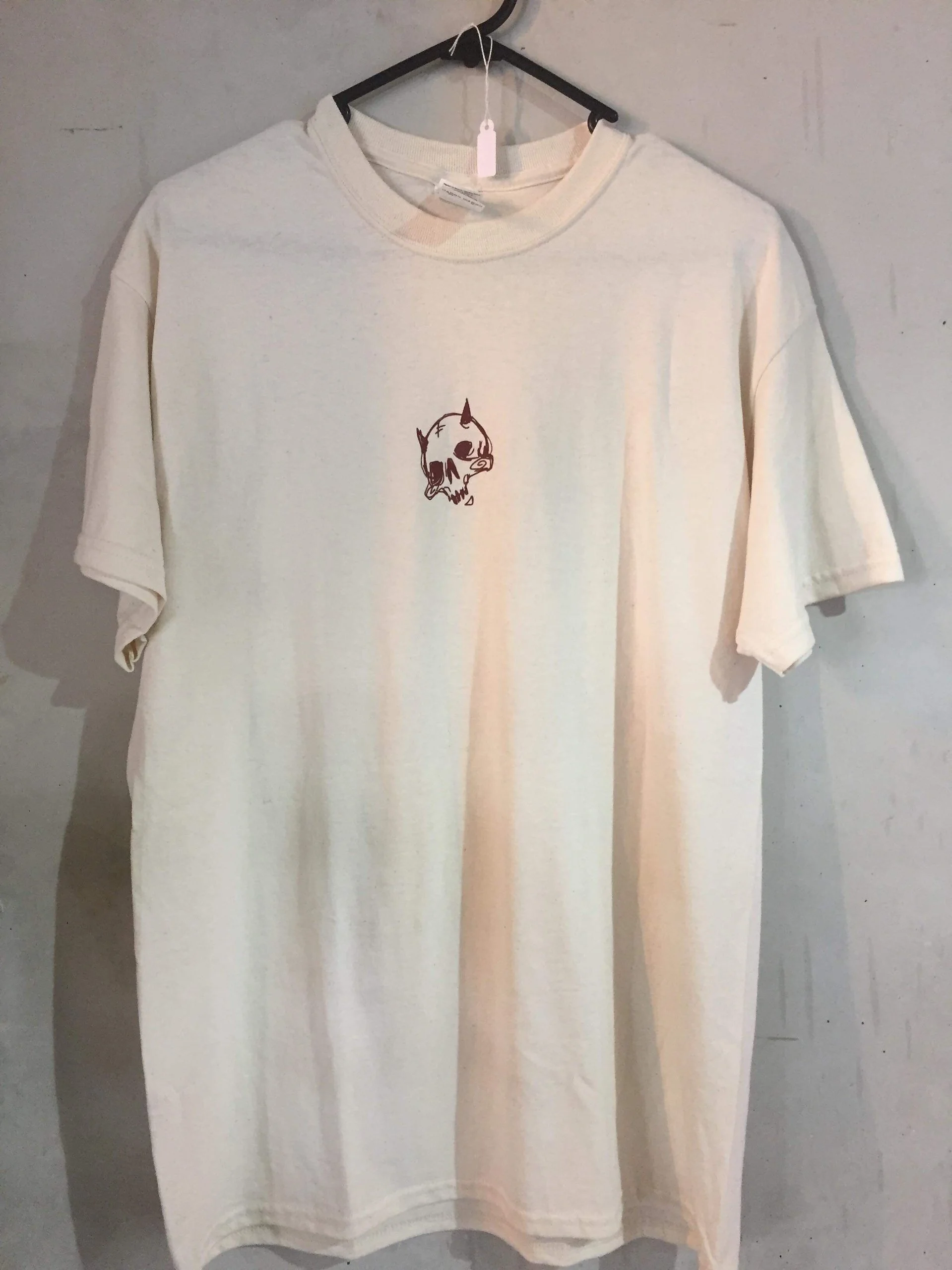 Cream-colored T-shirt hanging on a black hanger against a gray wall, with a small illustration of a cartoon dog's head with a skull face in the center.