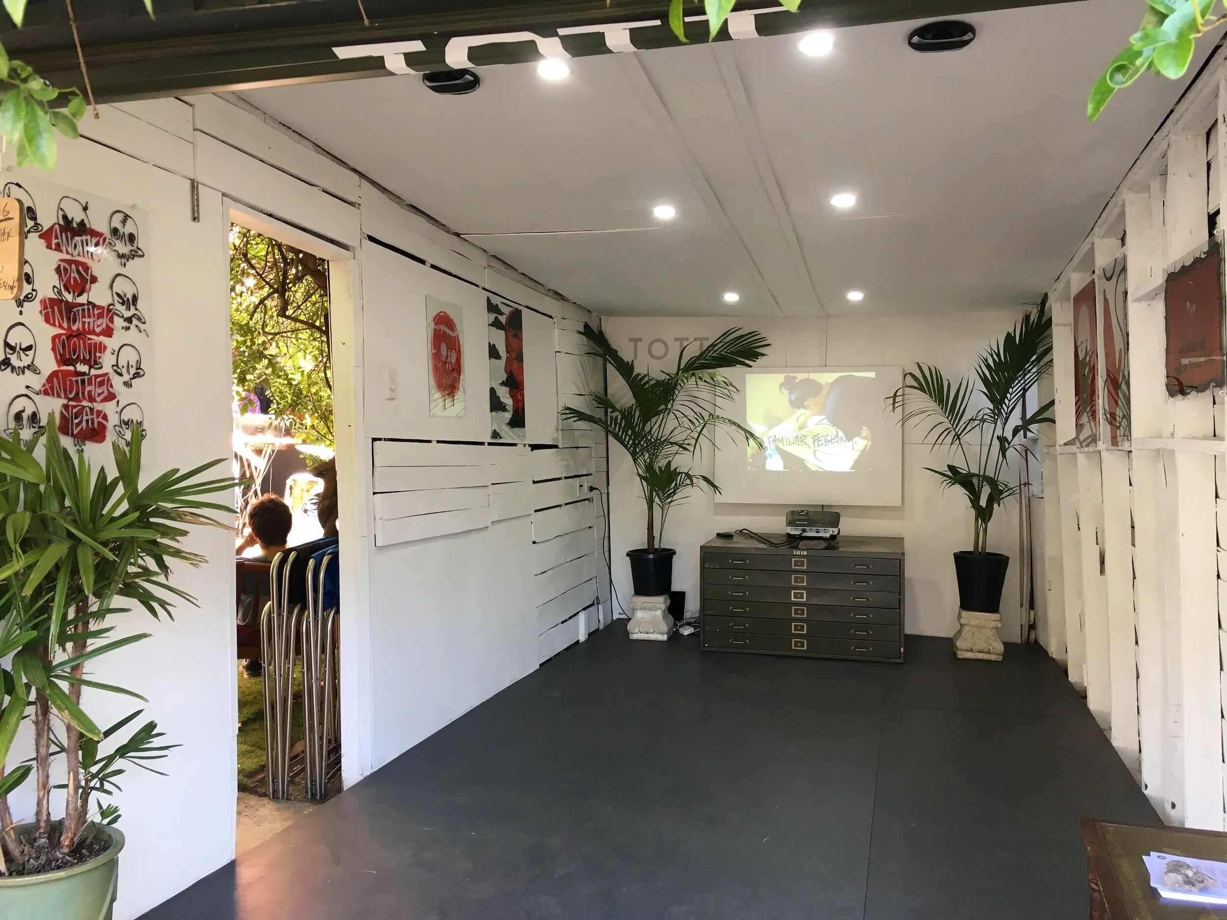 Indoor room with white walls decorated with art prints, potted plants, a projector screen showing a cartoon image, and a black file cabinet with drawers.