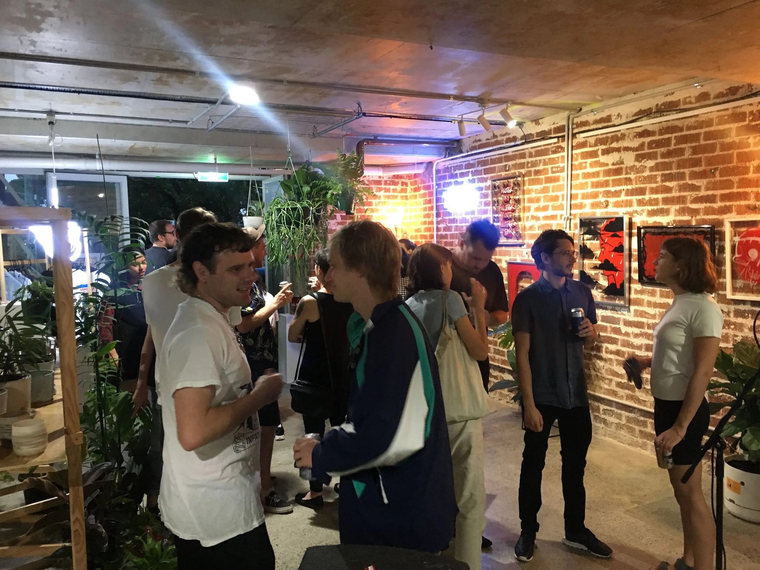 People socializing at an art gallery or event with brick walls and artwork displayed on the wall, indoor plant decorations, and warm lighting.