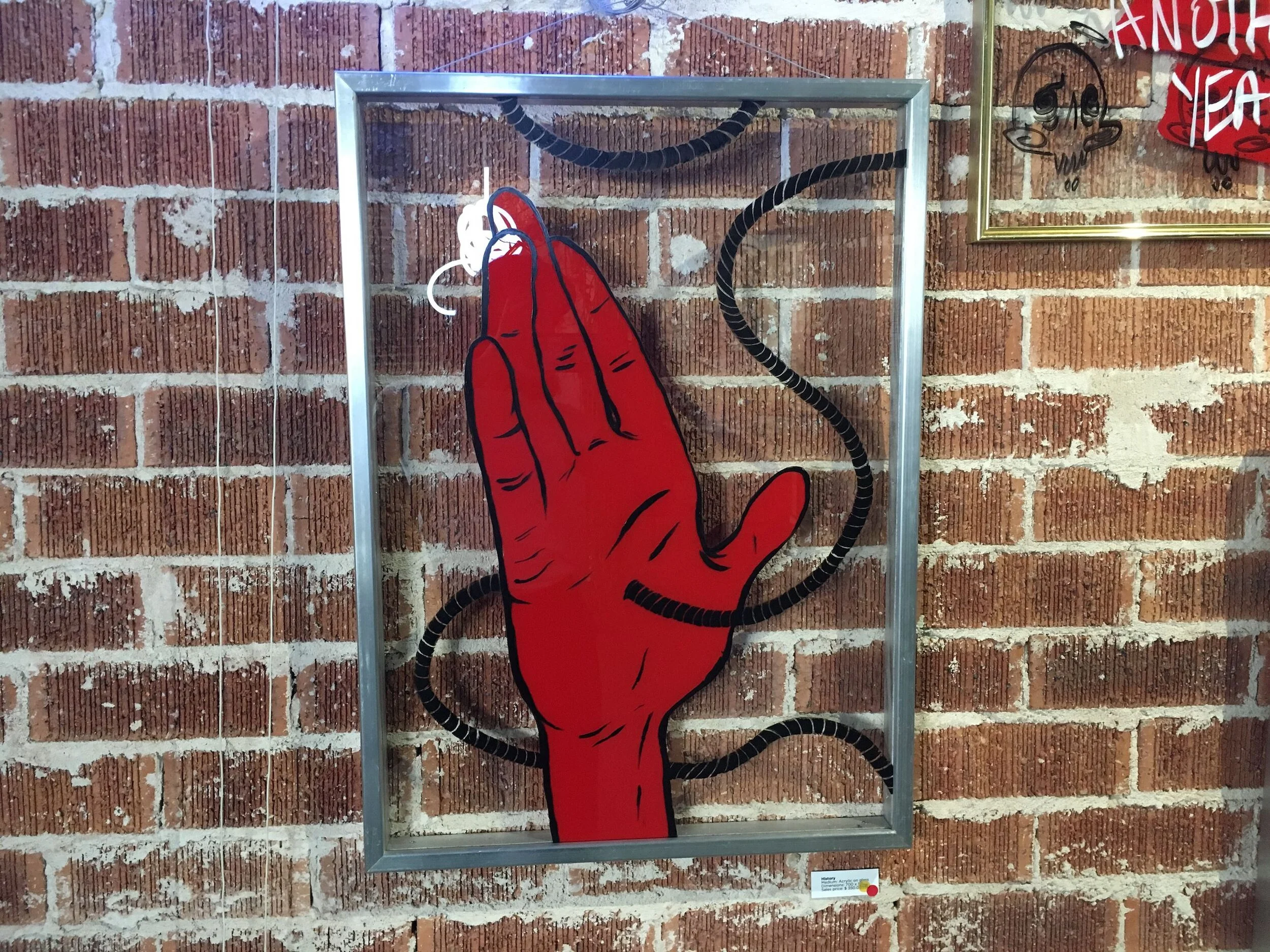 A framed pop art-style artwork of a red hand with black outlines on a brick wall, with a black cord looping around the hand and extending outside the frame.