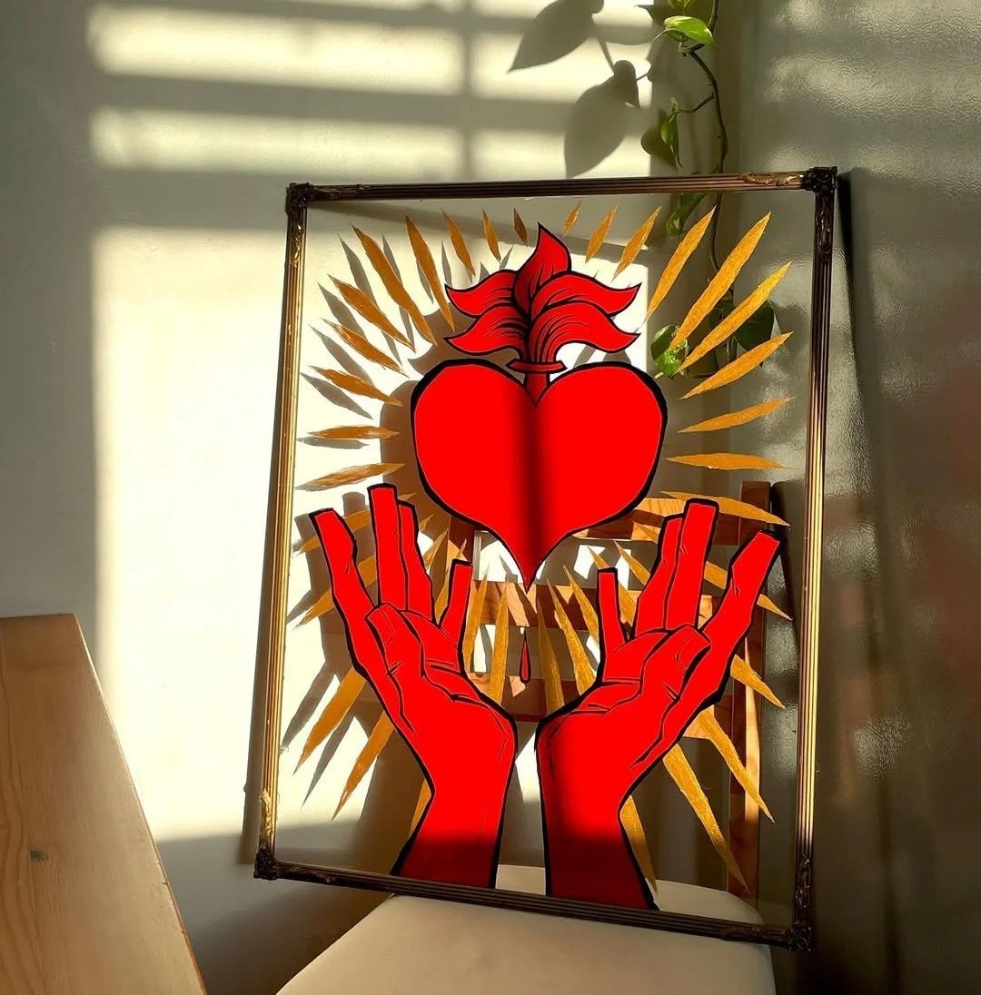 A piece of art featuring two red hands holding a red heart with a fiery top, with gold spikes radiating outward behind it, placed in a frame leaning against a wall with sunlight and shadow patterns.