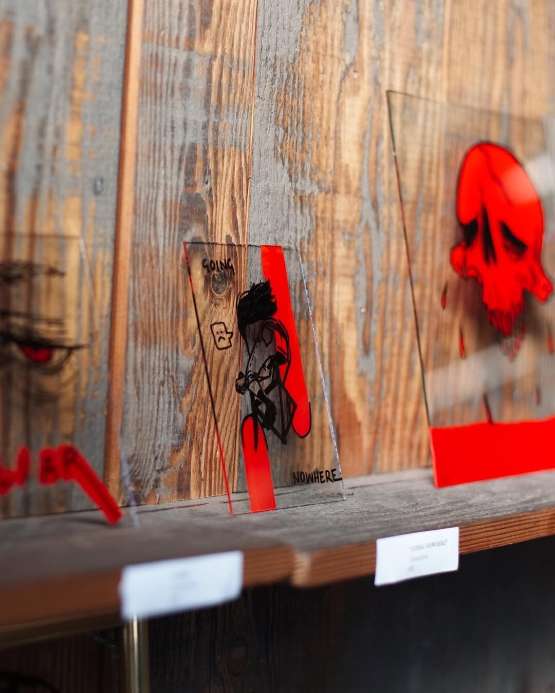 A clear acrylic sign featuring a stylized illustration of a person with long hair holding a phone or camera, with the words 'GOING NO WHERE' and a sad face, displayed on a wooden surface.