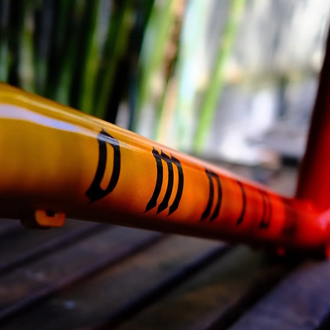 Close-up of a yellow bicycle frame with black markings, with blurred green plants and a window in the background.