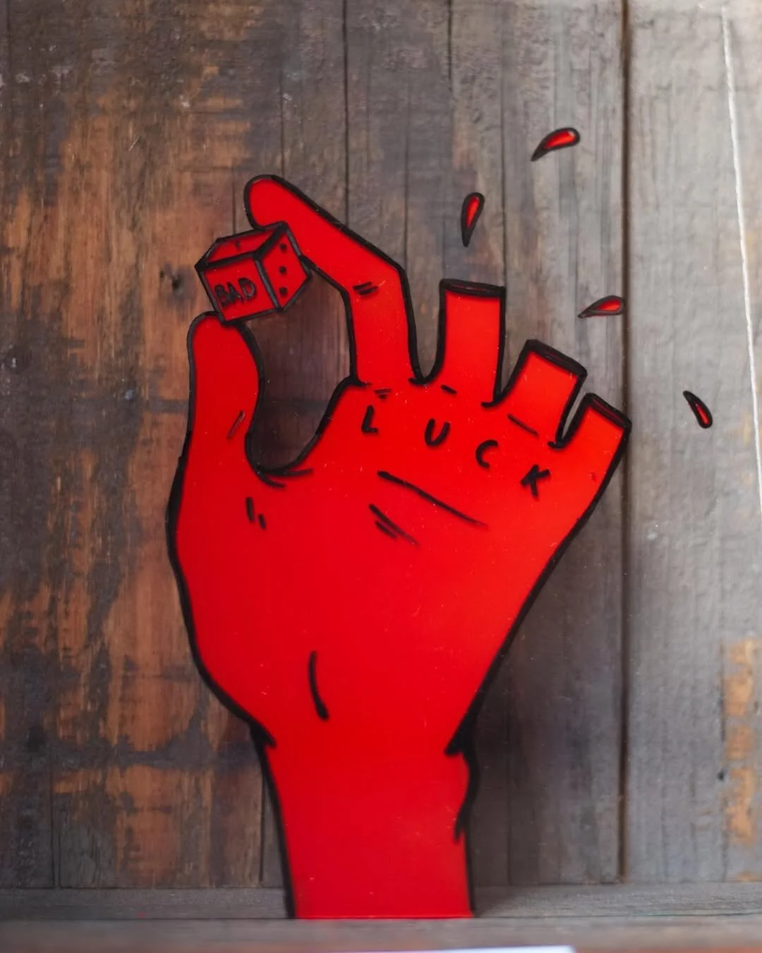 A red-colored artwork of a hand holding a small cube with the word 'BAD' written on it. The fingers of the hand have words written on them, and there are small red droplets and splashes around the art.