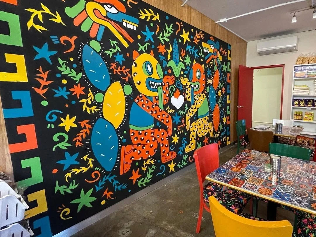 A colorful mural featuring cartoonish, abstract animal and humanoid figures, surrounded by vibrant flowers and foliage, with a decorative border on the left side.