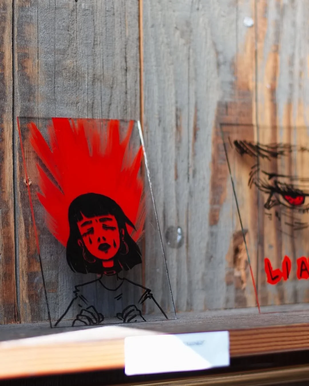A clear acrylic sign with a black and red illustration of a woman with short hair, her eyes closed and lips parted, with red streaks radiating from her head, placed against a wooden wall with other artwork visible in the background.