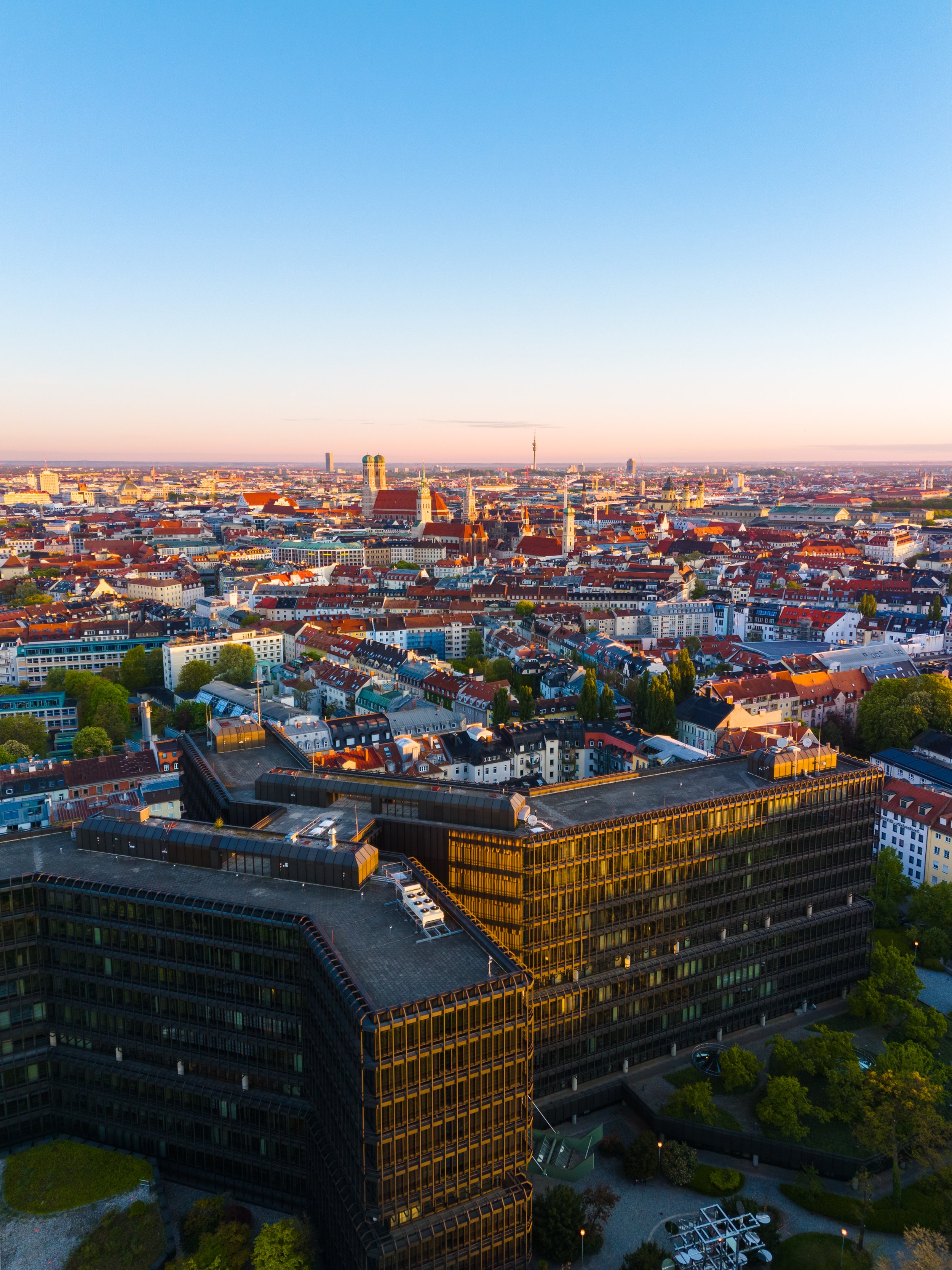 Luftaufnahme von München bei Sonnenuntergang, mit bekannten Bauwerken und einem modernen, großen Gebäude im Vordergrund. Der Himmel ist klar und blau, während der Horizont ein warmes Licht hat.