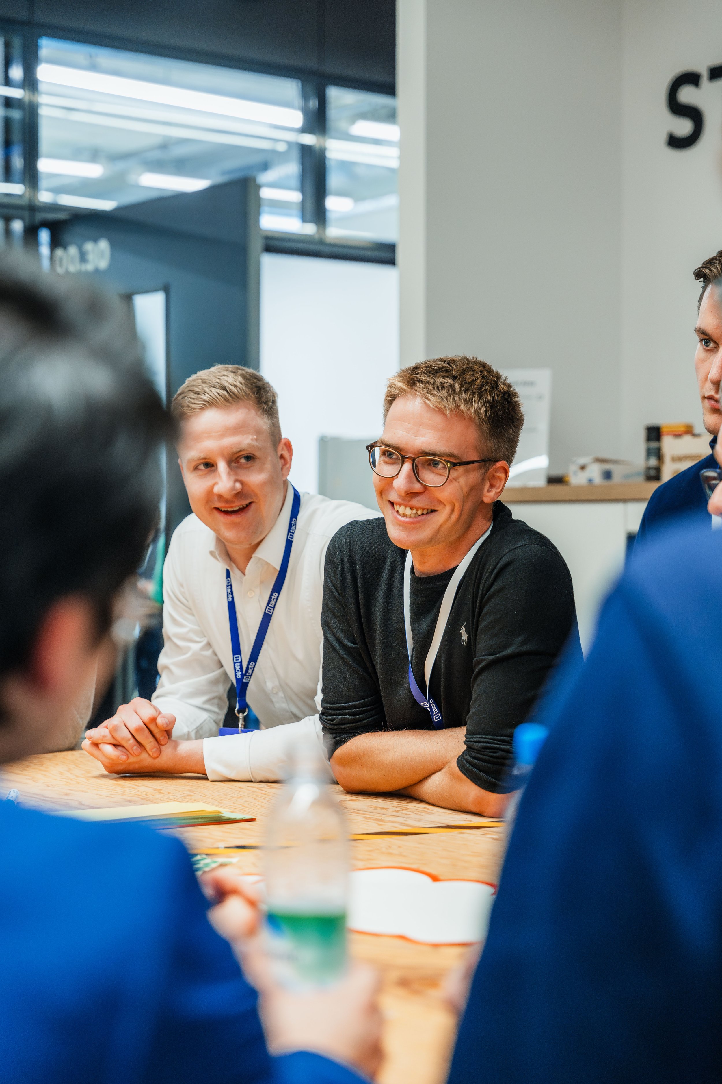 Gruppe von Personen in einem Büro bei einer Besprechung am Tisch sitzend, zwei Männer lächeln, beide tragen ID-Badges.