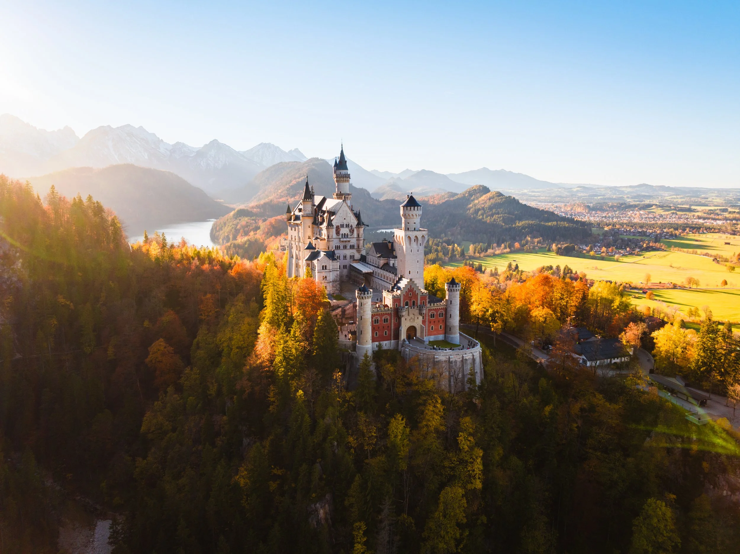 Luftaufnahme von Schloss Neuschwanstein in Deutschland im Herbst, umgeben von bunten Wäldern und Bergen im Hintergrund.