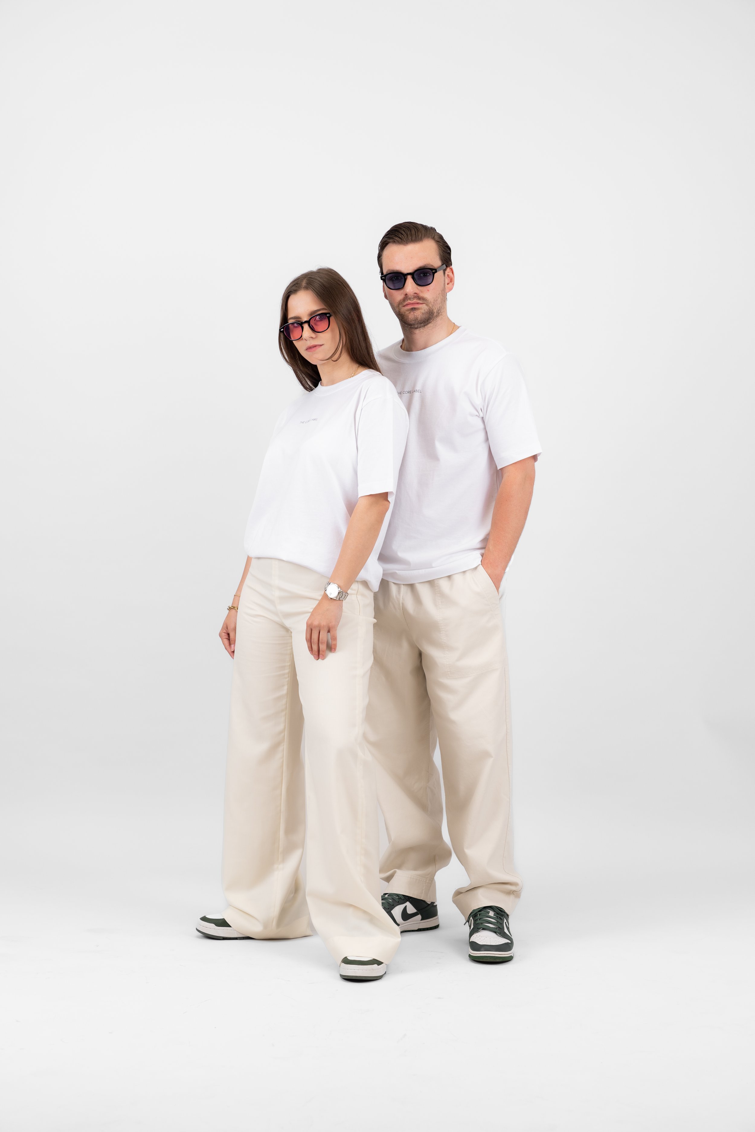 Ein Mann und eine Frau in lässiger Kleidung stehen nebeneinander. Beide tragen weiße T-Shirts und beigefarbene Hosen. Sie haben Sonnenbrillen auf und posieren vor einem weißen Hintergrund.
