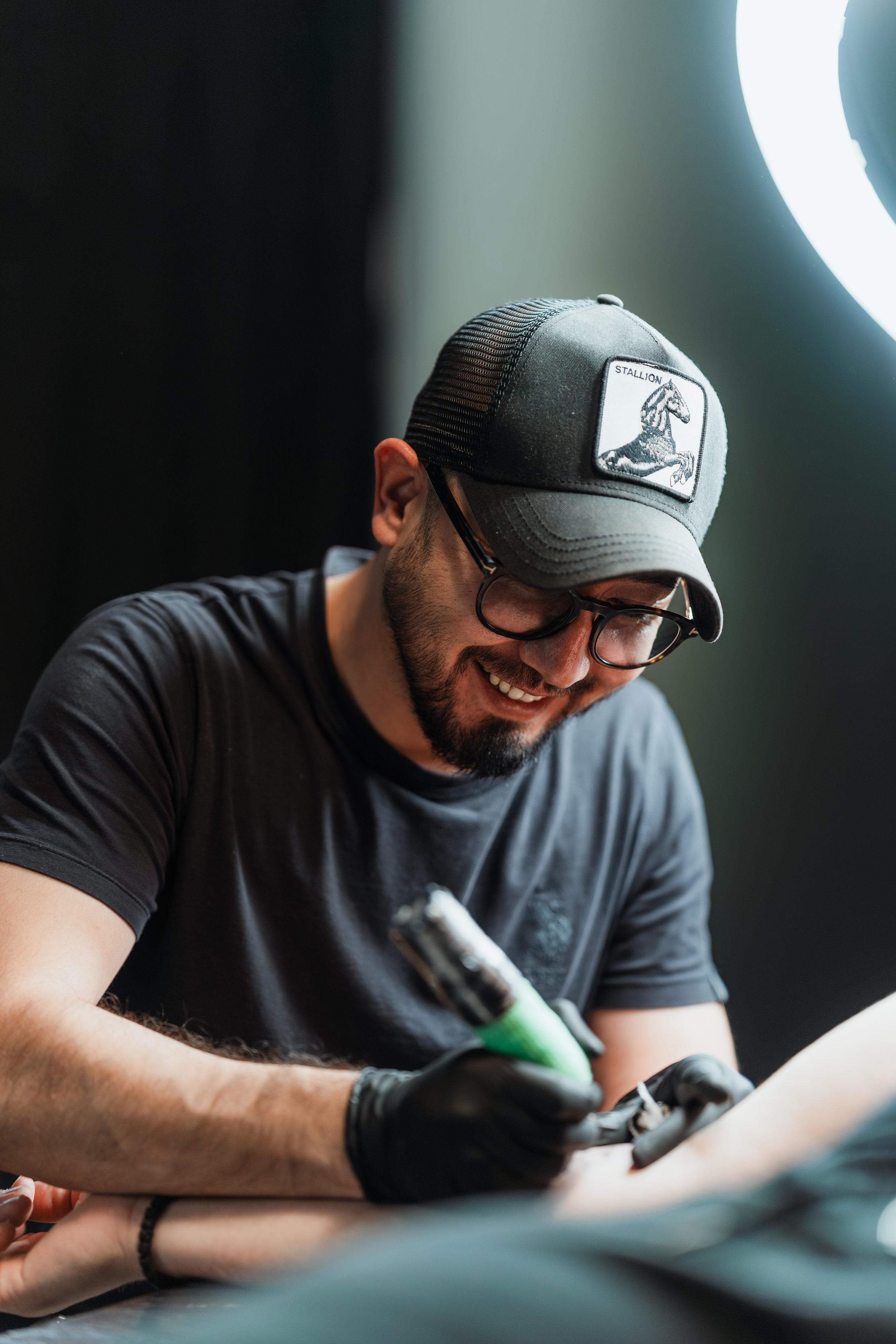 Ein Mann mit Bart und Brille trägt eine schwarze Baseballkappe und tätowiert jemanden. Er lächelt und hat schwarze Handschuhe an. Der Hintergrund ist unscharf und dunkel.