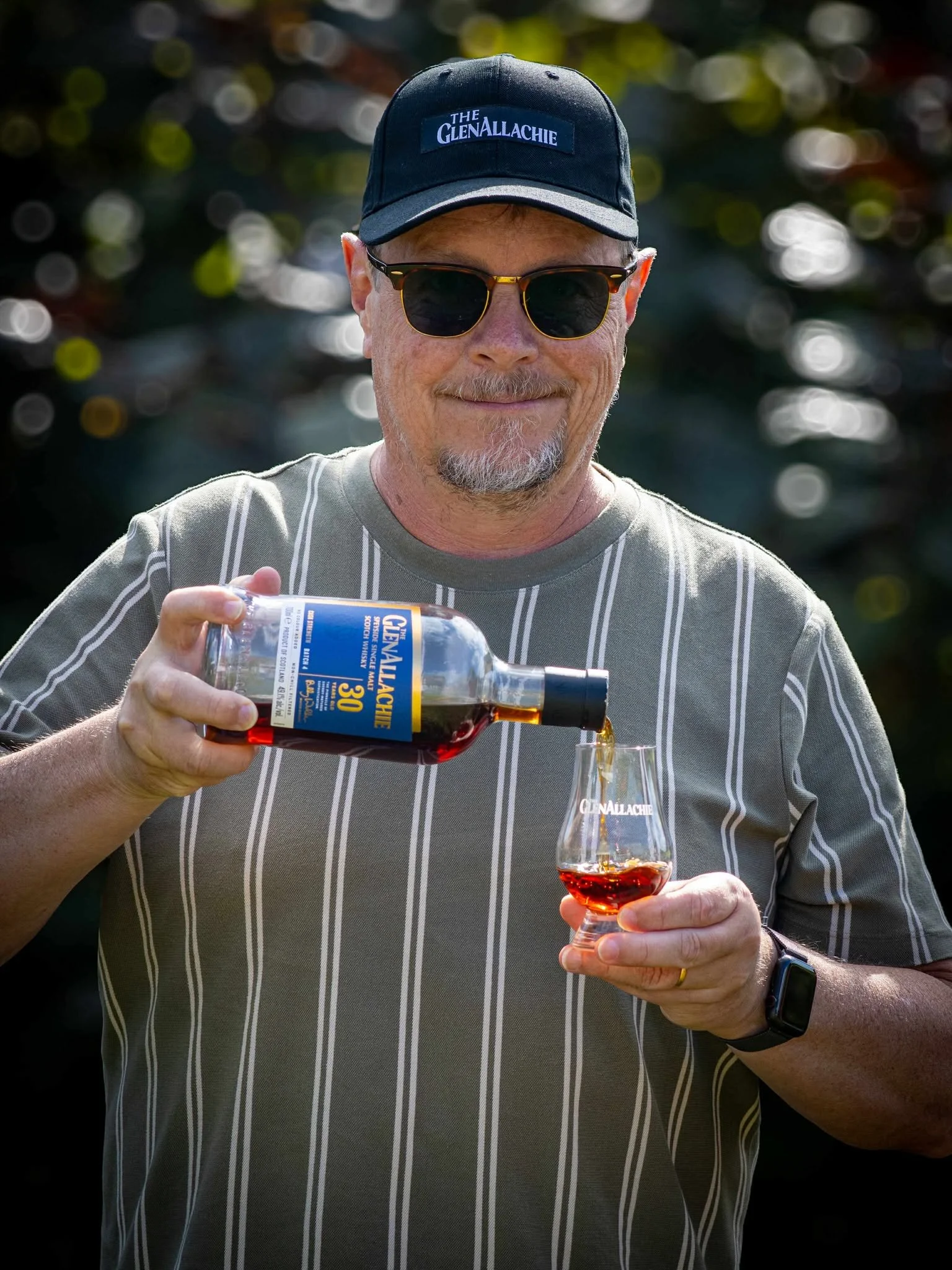 A man standing outdoors wearing a black T-shirt and a black cap, with trees and a wooden fence in the background.