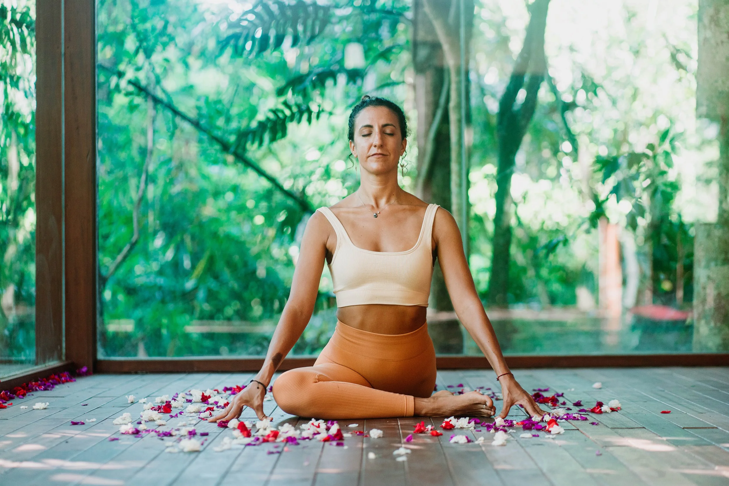 A woman practicing yoga outdoors on wooden decking, sitting cross-legged with eyes closed, surrounded by flower petals, with lush green trees in the background.