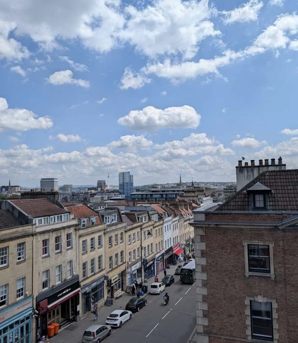 View of a city street with historic buildings, parked cars, and people walking, under a blue sky with scattered clouds.
