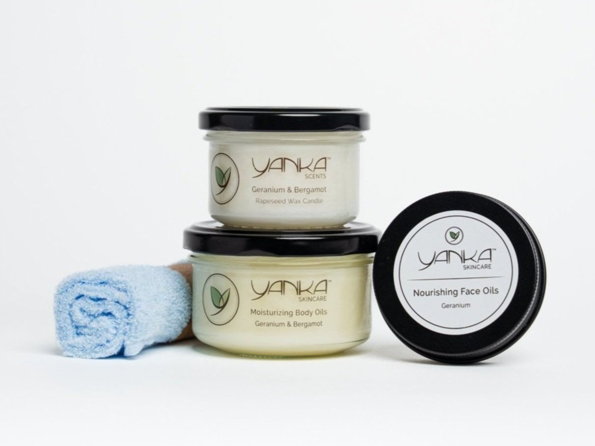 Yanka Skincare products with geranium & bergamot, including a candle, body oil, and face oil, arranged with a blue facecloth on a white background.