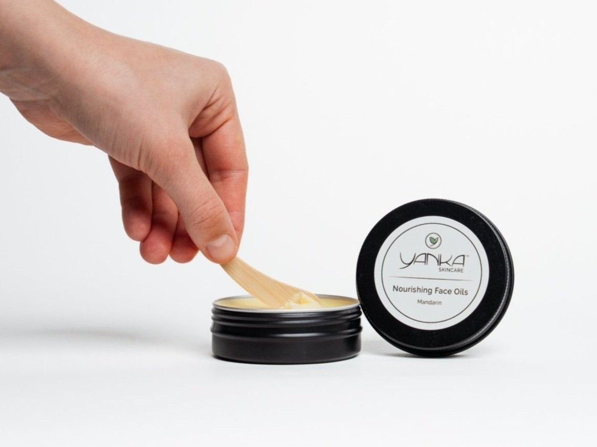 A hand using a wooden spatula to scoop balm from an open tin of Yanka Skincare Nourishing Face Oils, with the lid beside it on a white background.