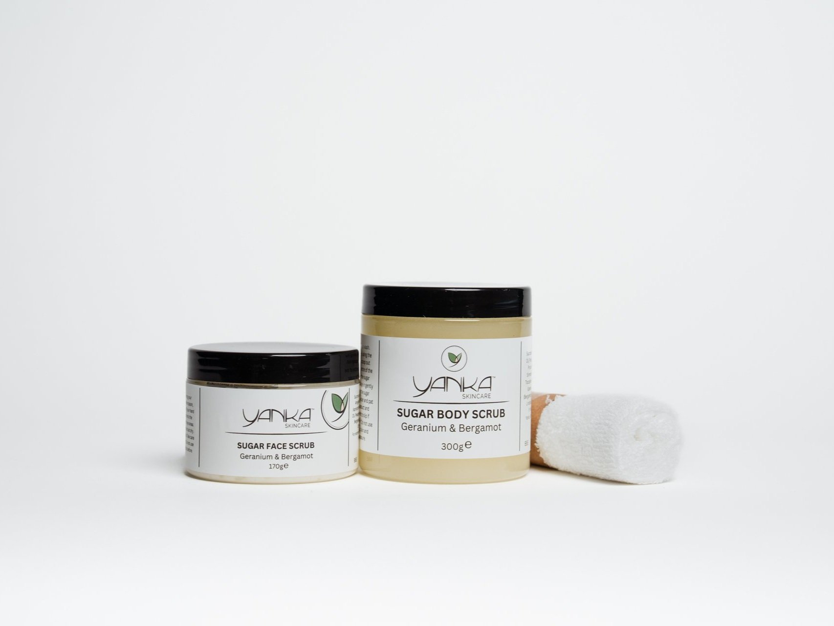 Yanka Skincare Sugar Face Scrub and Sugar Body Scrub in Geranium & Bergamot scent, placed next to a rolled white facecloth on a white background.