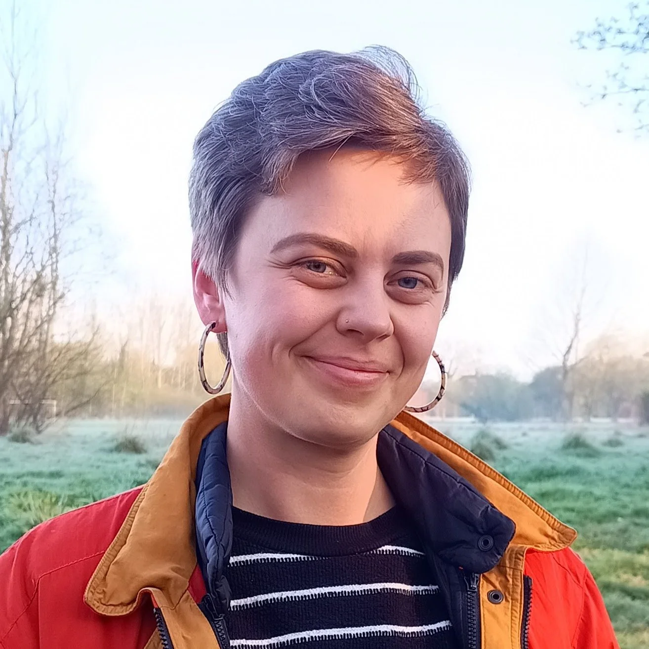 A photo of Kate Parkins smiling. She's wearing a red coat and a black/white striped jumper. Grass and trees can be seen in the background.