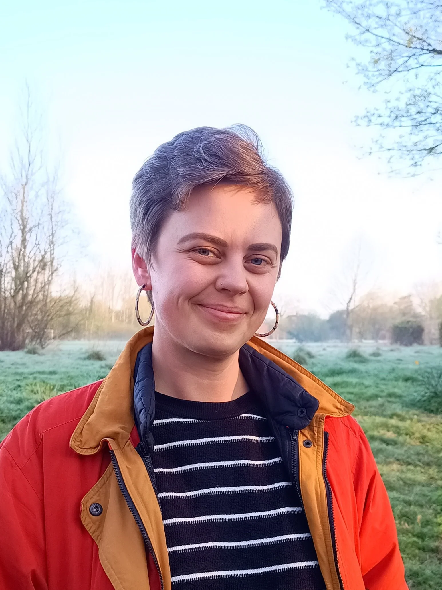 A photo of me, Kate Parkins, smiling and stood in front of a field and trees. I'm wearing a black and white striped jumper and a red coat.