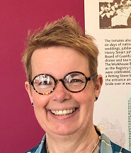A woman with short blonde hair, glasses, and a bright smile standing against a pink wall.