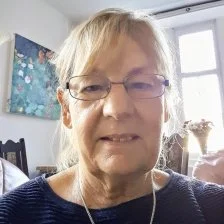 portrait of an older woman with blond hair and glasses, smiling indoors with a window and painting in the background.