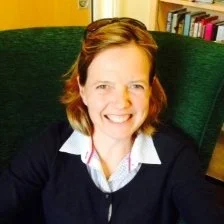 A woman with short red hair and fair skin smiling at the camera, sitting in a green armchair in a room with bookshelves.