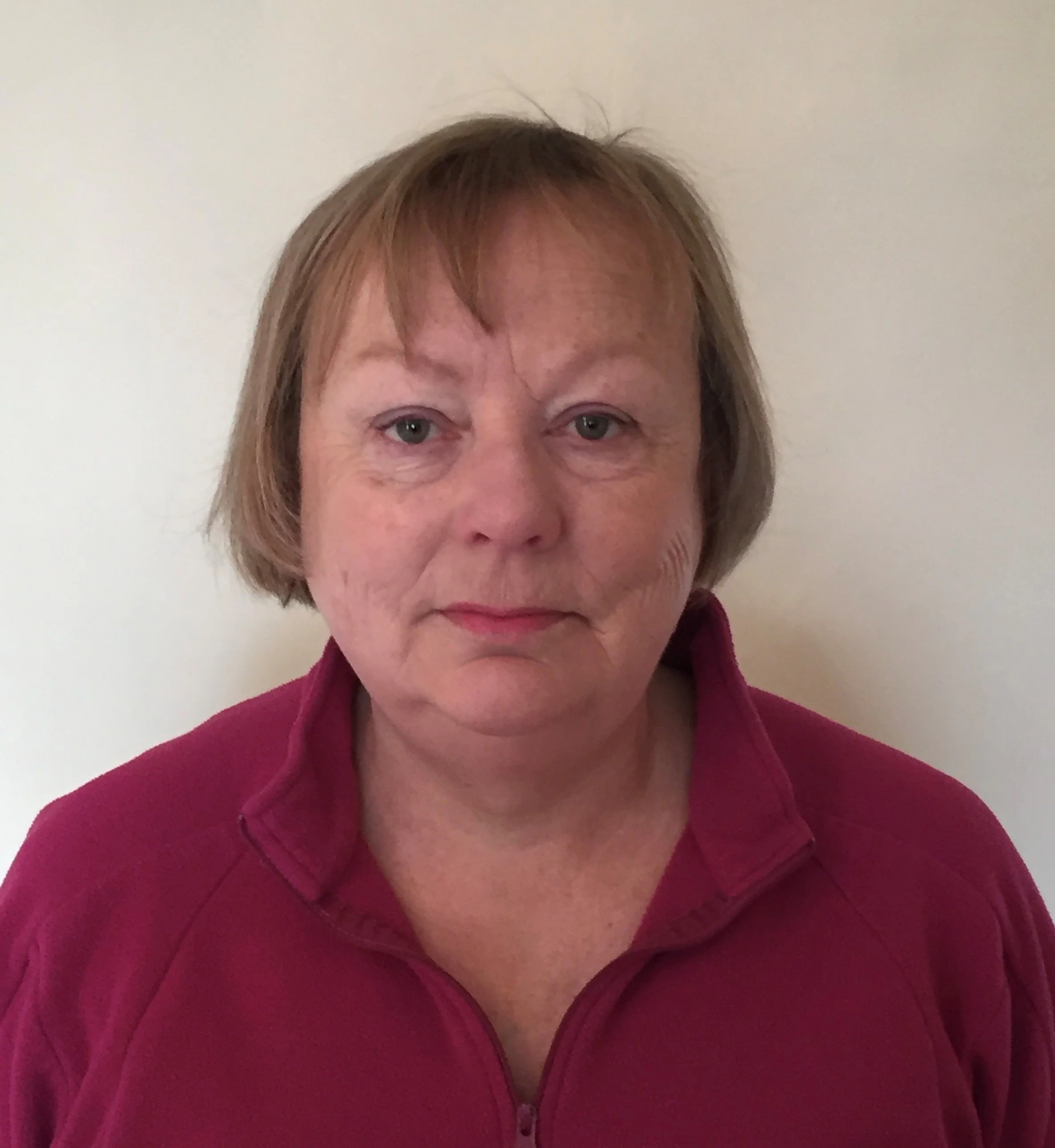 A middle-aged woman with short, light brown hair, wearing a maroon zip-up jacket, standing against a plain white wall.