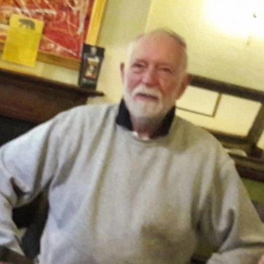 An elderly man with white hair and a beard sitting indoors, smiling, wearing a light gray sweatshirt with a black collar, in a room with yellow walls and framed pictures.