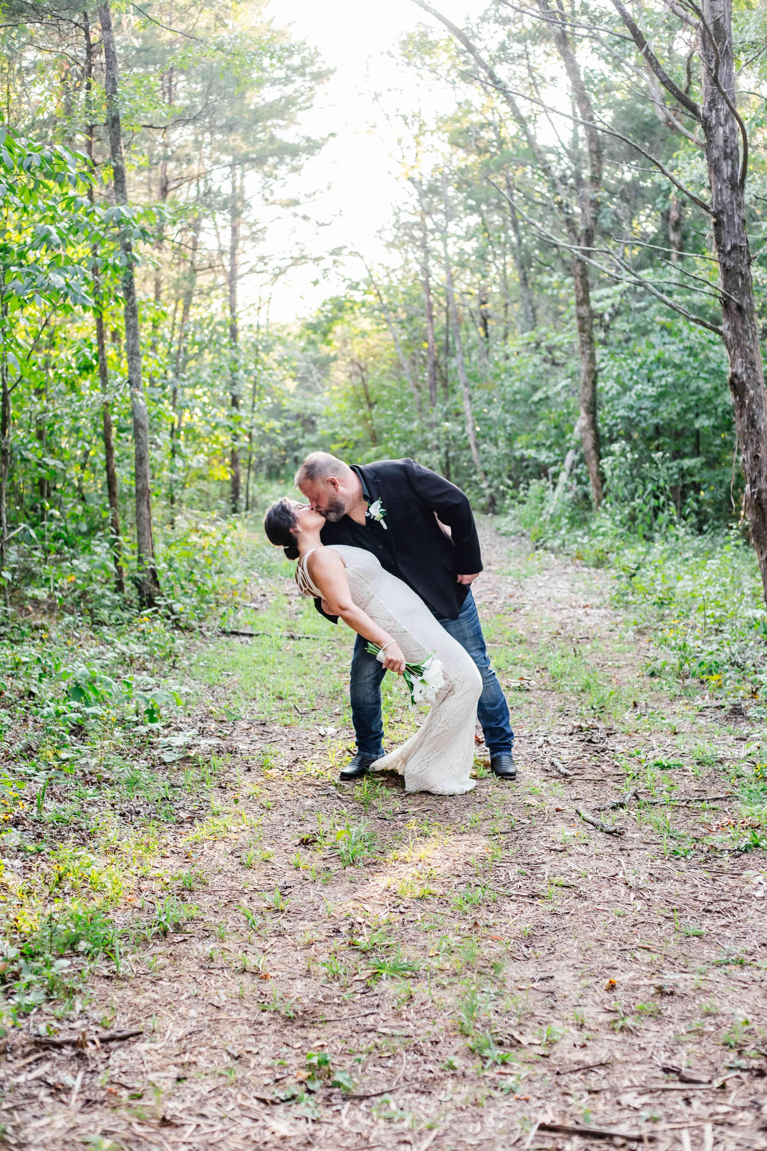 A couple in wedding attire sharing a kiss in a forested area during the daytime, with green trees and sunlight in the background.