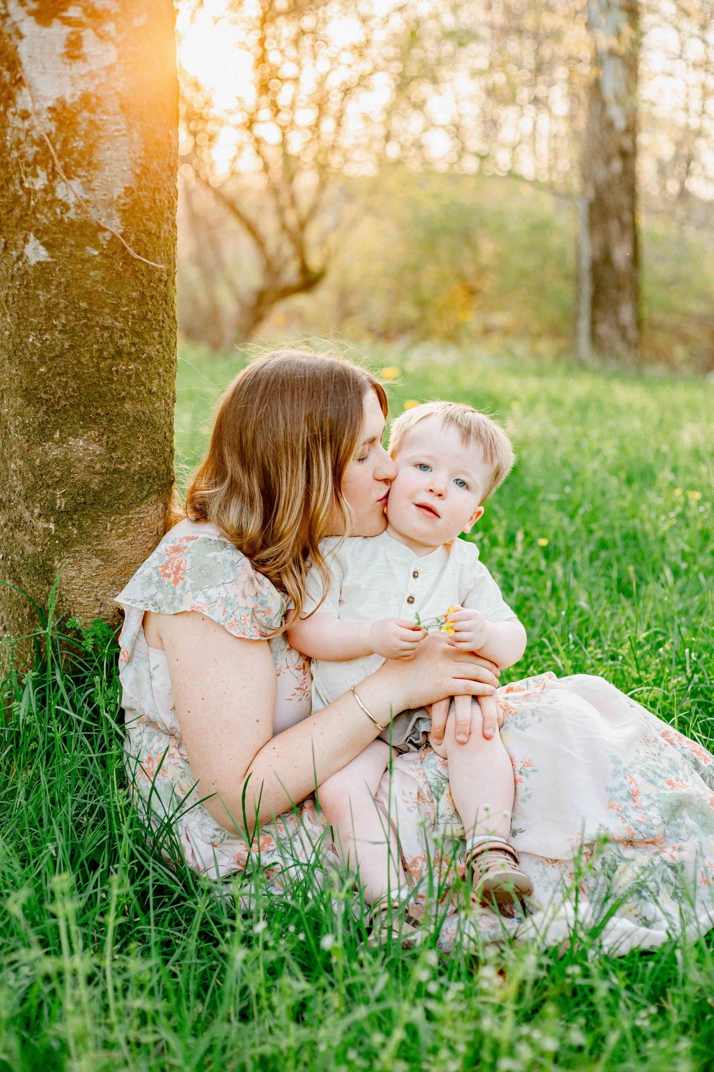 A woman sitting outdoors against a tree, kissing a young boy on the cheek during sunlight, with green grass and trees in the background.