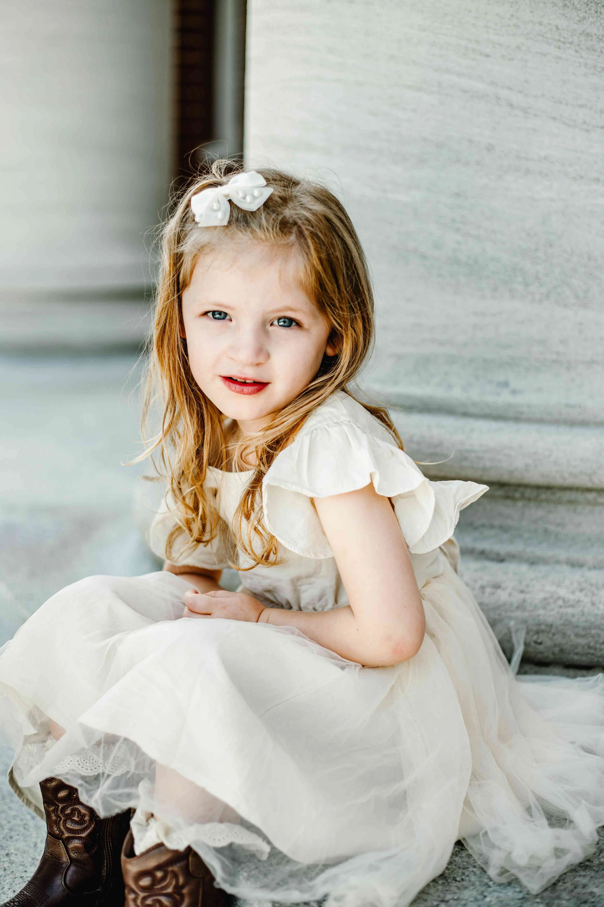 A young girl with red hair, blue eyes, and a white bow in her hair, sitting on the ground near a building, wearing a light-colored dress and brown boots, looking at the camera.