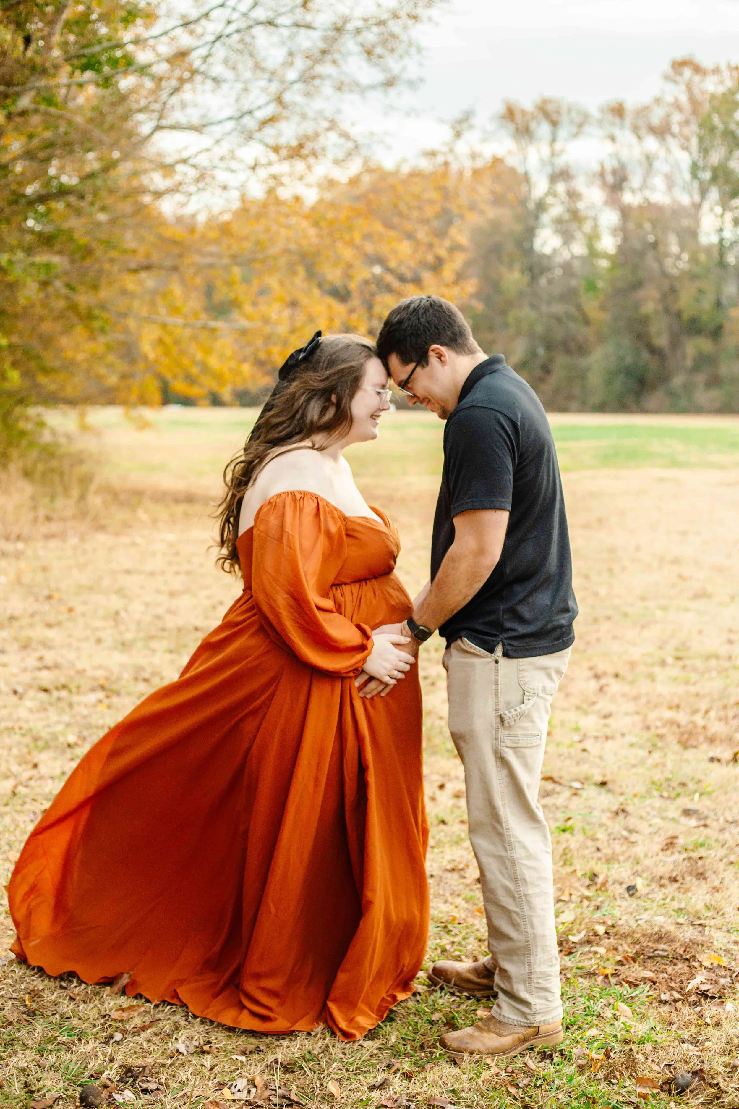 A couple with light skin standing outdoors in fall, holding hands, with their foreheads touching, smiling, surrounded by trees with autumn leaves.