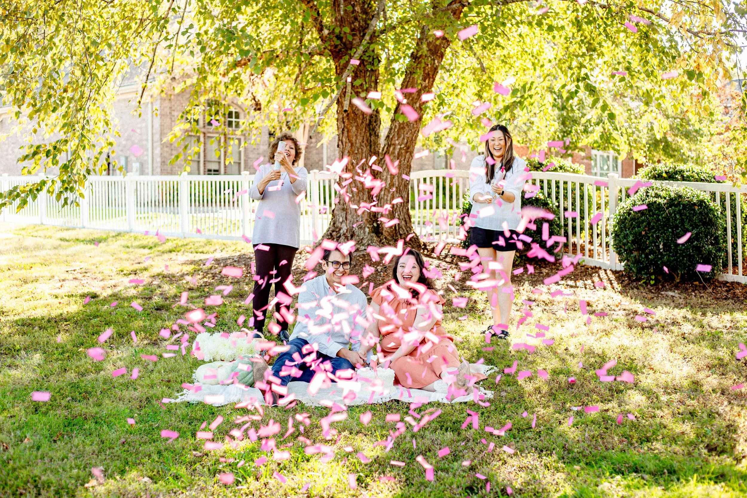 Group of four friends celebrating outdoors under a large tree, surrounded by greenery and a white picket fence. Two are sitting on a blanket with a cake, while two are standing and popping confetti into the air, all smiling and joyful.