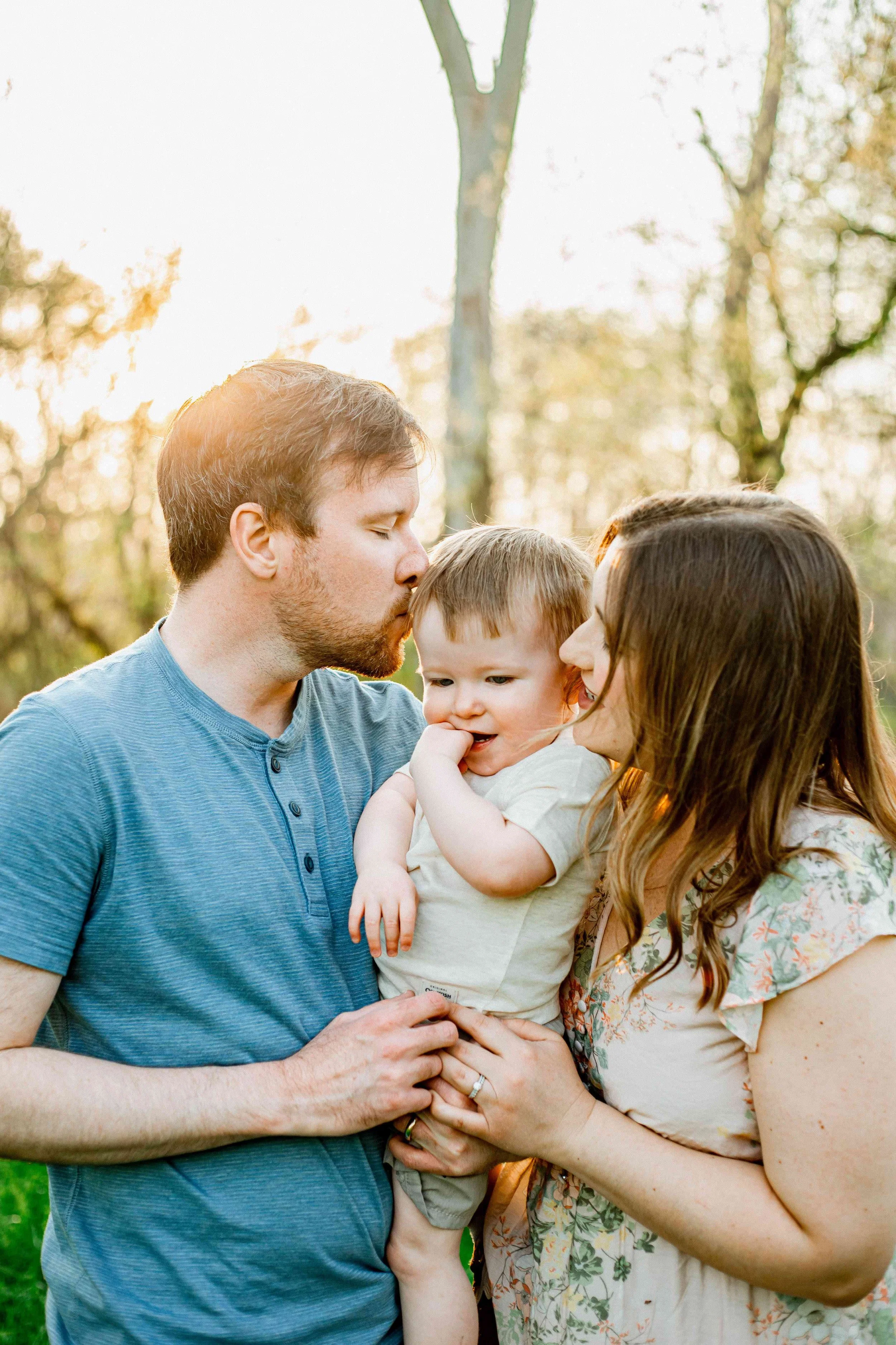 A family of three, with a father, mother, and their young son, outside in a park during sunset, sharing a tender moment.