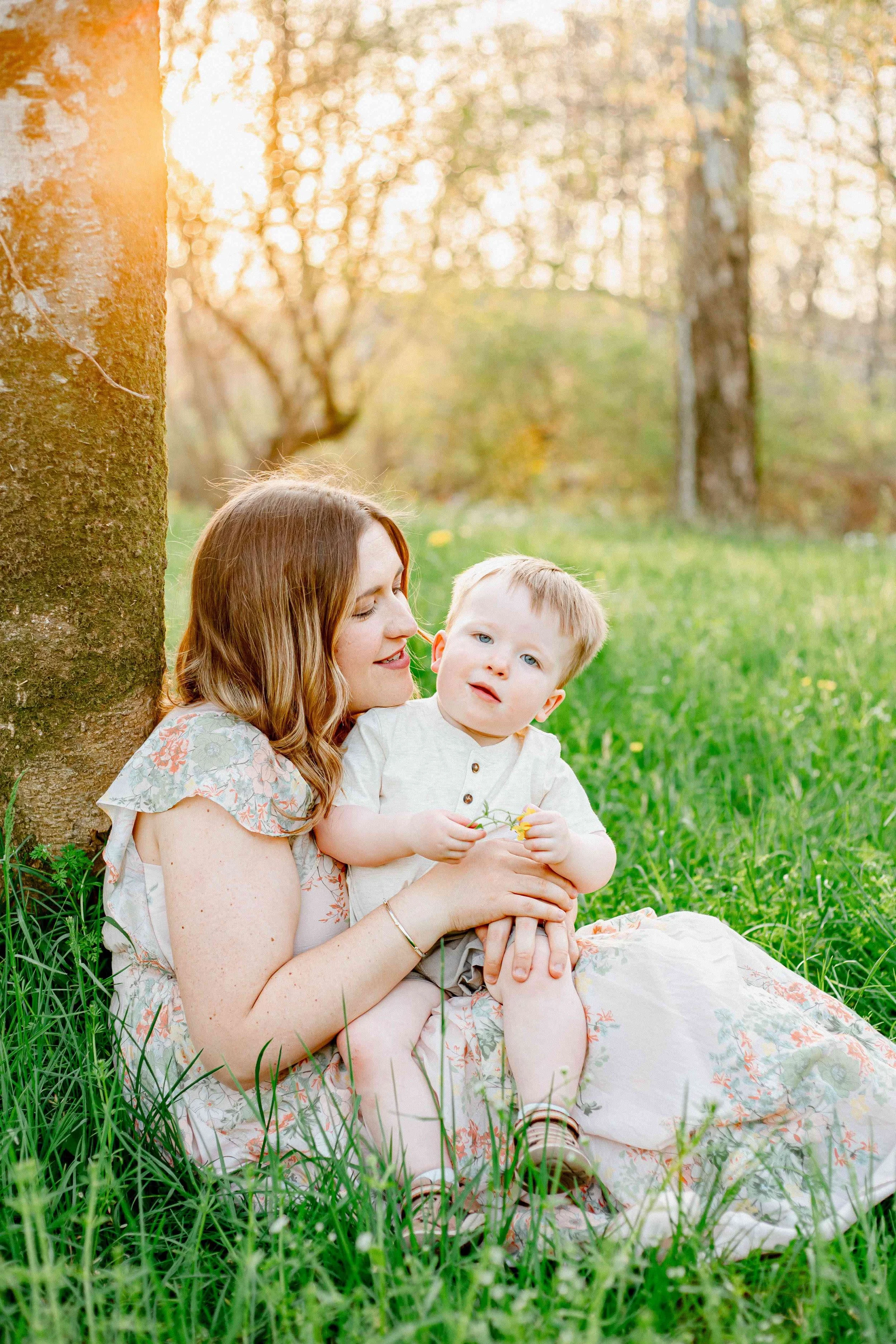 A woman sitting on the grass leaning against a tree trunk, holding a young boy in her lap, outdoors in a sunlit park during spring or summer, with green grass and trees in the background.