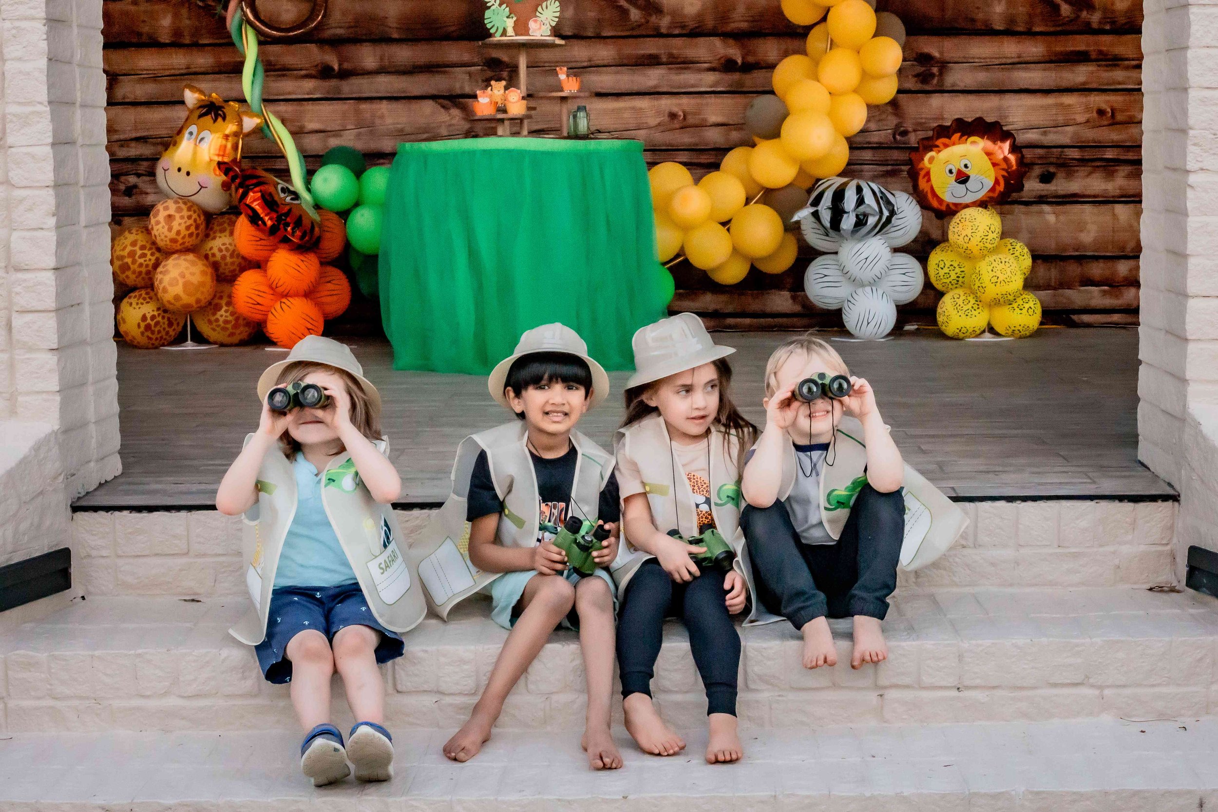 Four children sit on a fireplace step during a jungle-themed birthday party, wearing safari hats and vests, with binoculars and small toy animals, while decorations like balloons and animal cutouts are in the background.