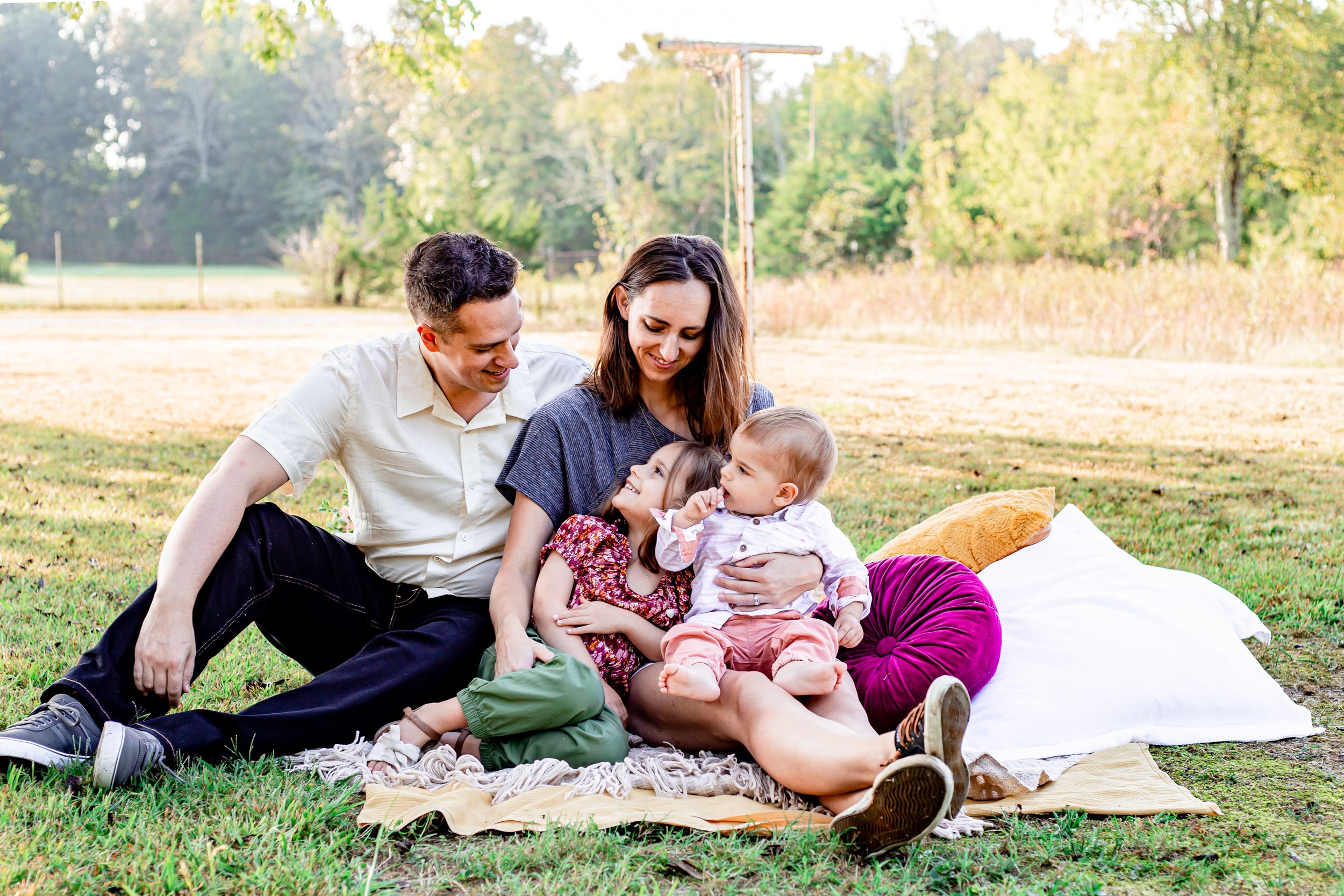 Family of four sitting on a blanket outdoors, smiling and enjoying each other's company in a grassy field with trees in the background.