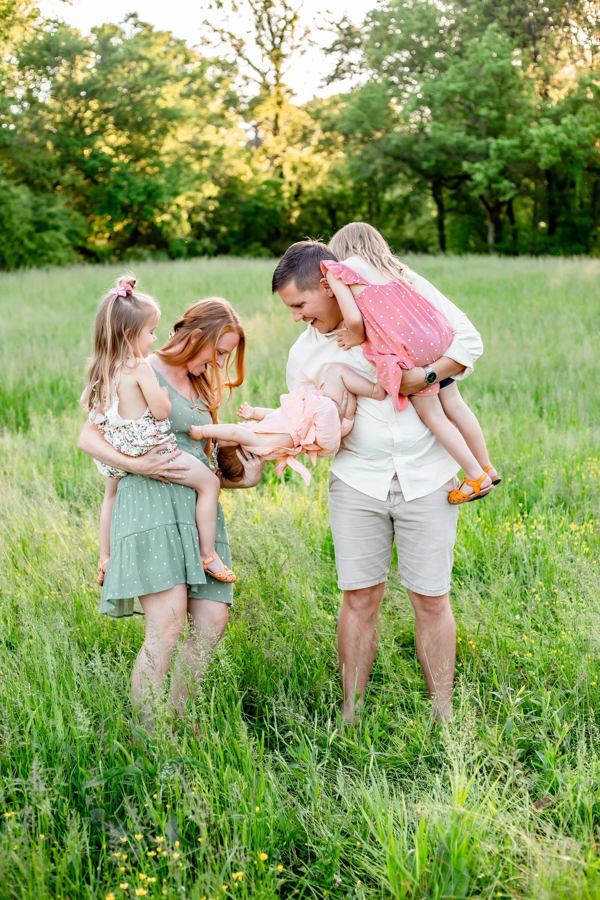 A family of five, including two young girls and two adults (a man and a woman), playing together outdoors in a grassy field with trees in the background. The man is holding one girl while the woman holds the other girl, and they are all smiling and laughing.