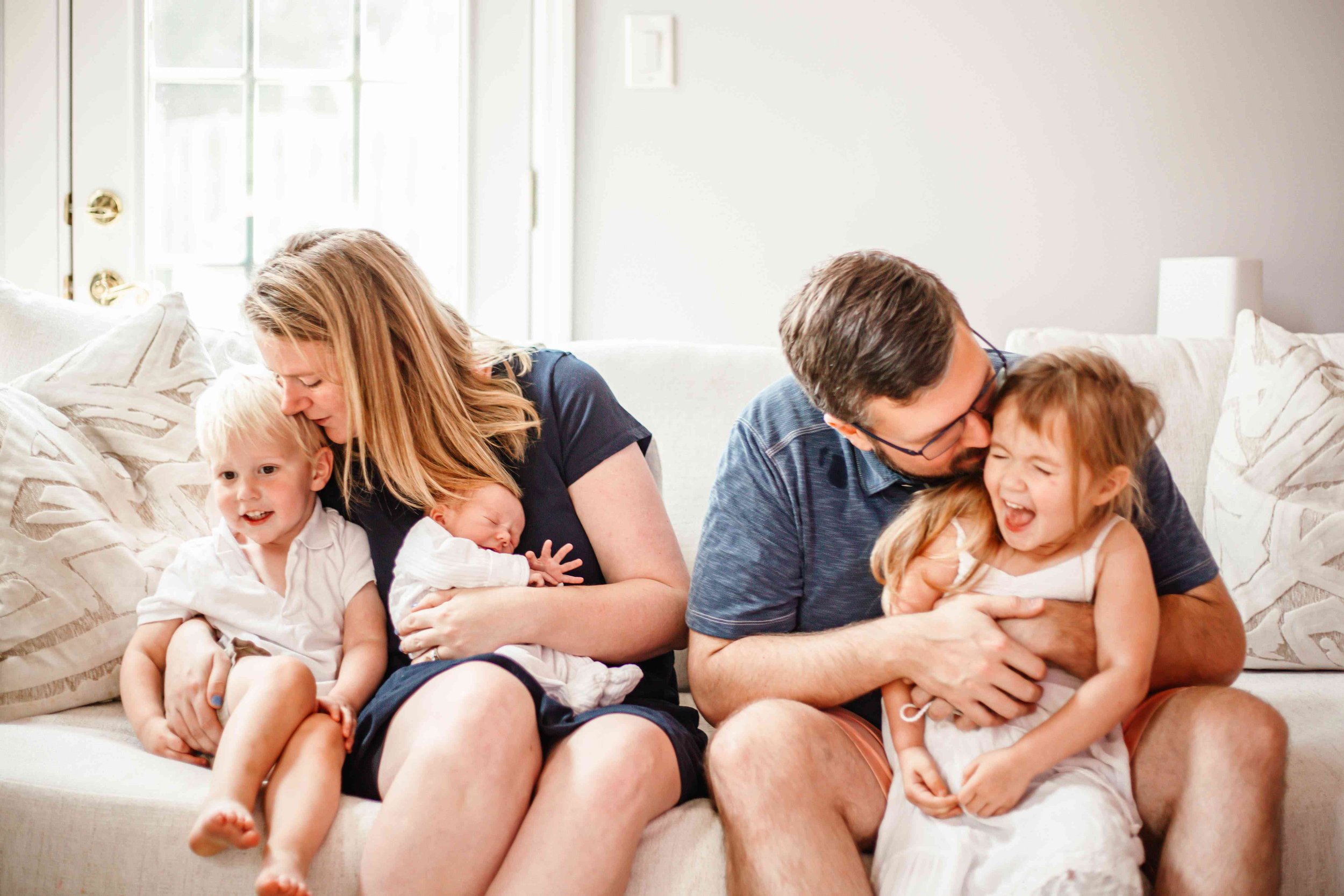 Family of two parents with four young children sitting on a white couch, embracing and all smiling happily in a bright living room.