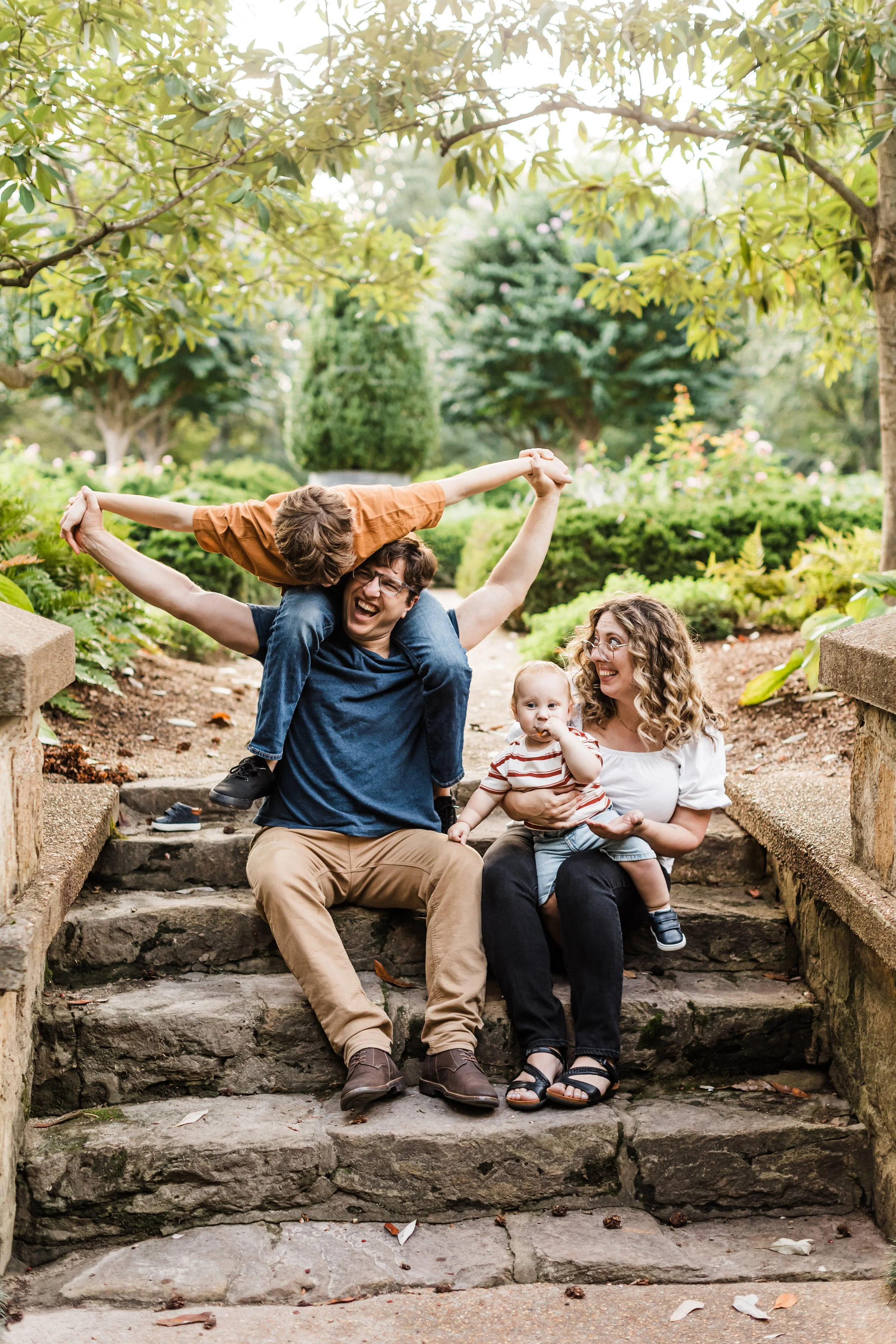 A family of four, including a father, mother, young boy, and baby, sitting on stone steps in a park with greenery, trees, and shrubs in the background, enjoying a playful moment together.