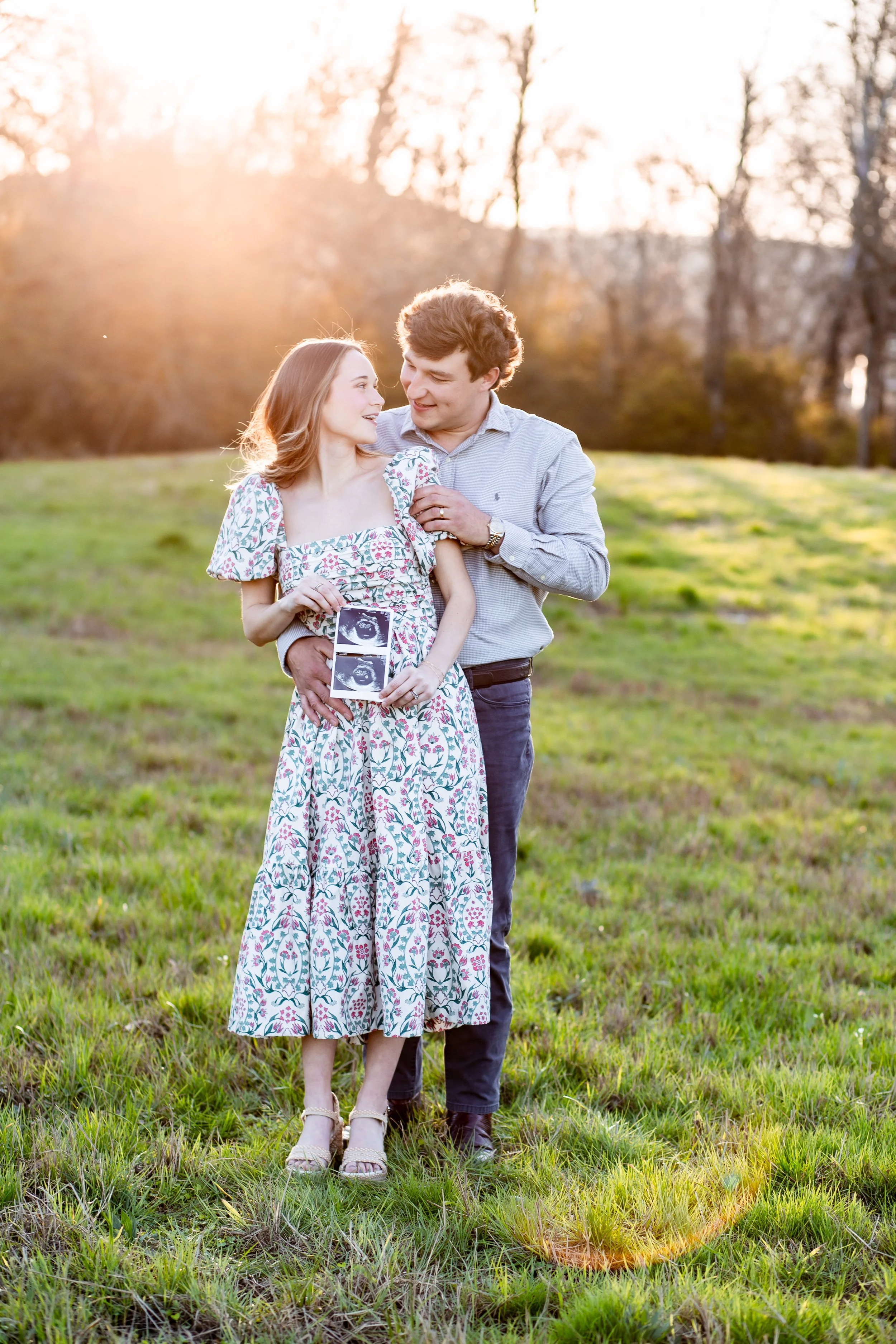 A couple standing outdoors in a grassy field at sunset, holding ultrasound images and celebrating a pregnancy.