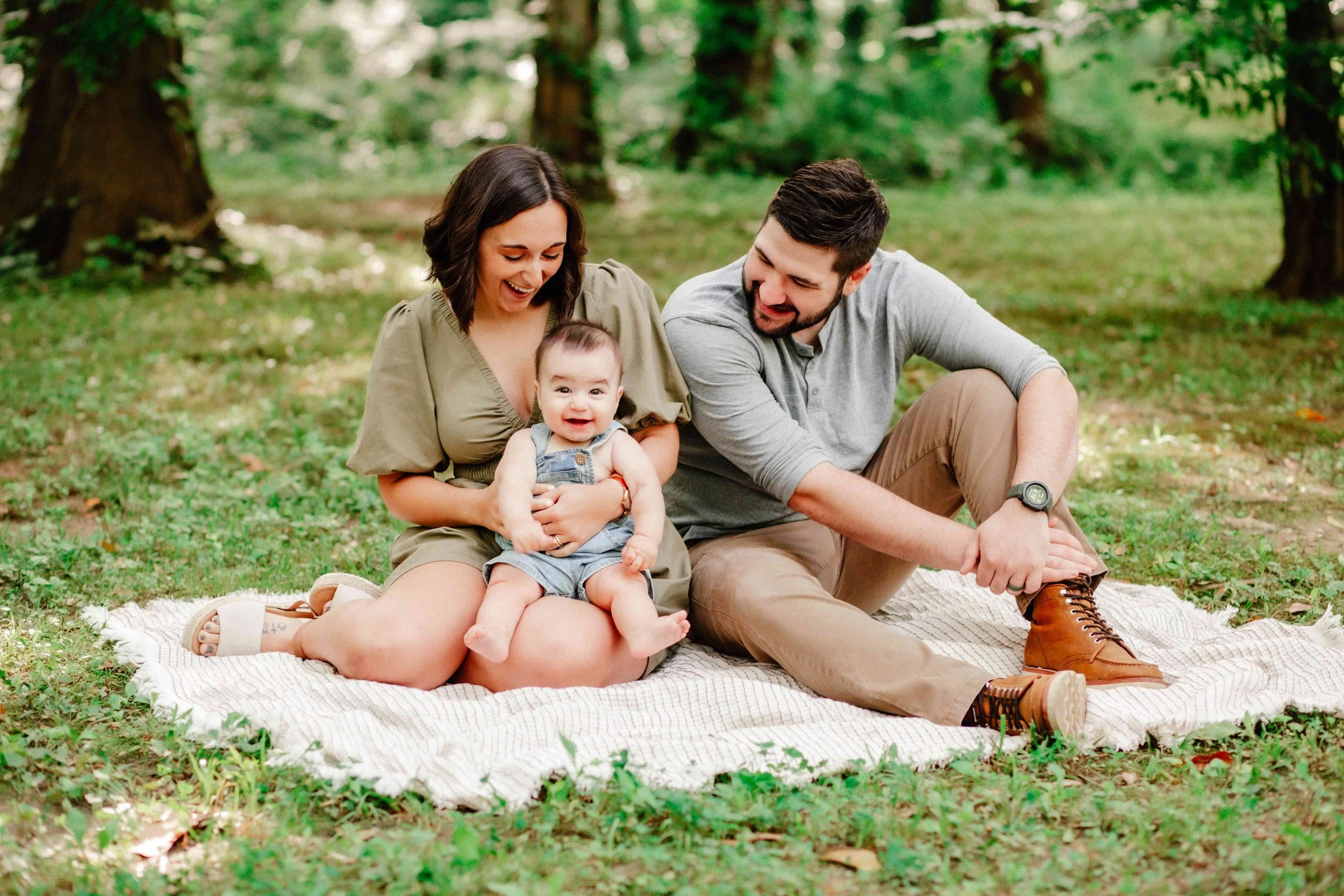 Family sitting on a blanket in a park, smiling and playing with a baby, surrounded by trees.