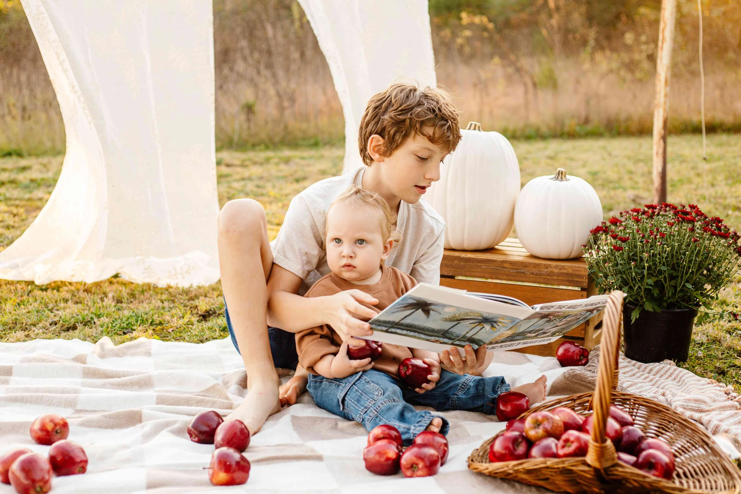 Two children, a boy and a toddler, sit on a picnic blanket outdoors, with apples, pumpkins, and flowers around them. The boy is reading a book to the toddler, who is holding an apple. There is a wooden crate with white pumpkins, and a potted plant nearby.